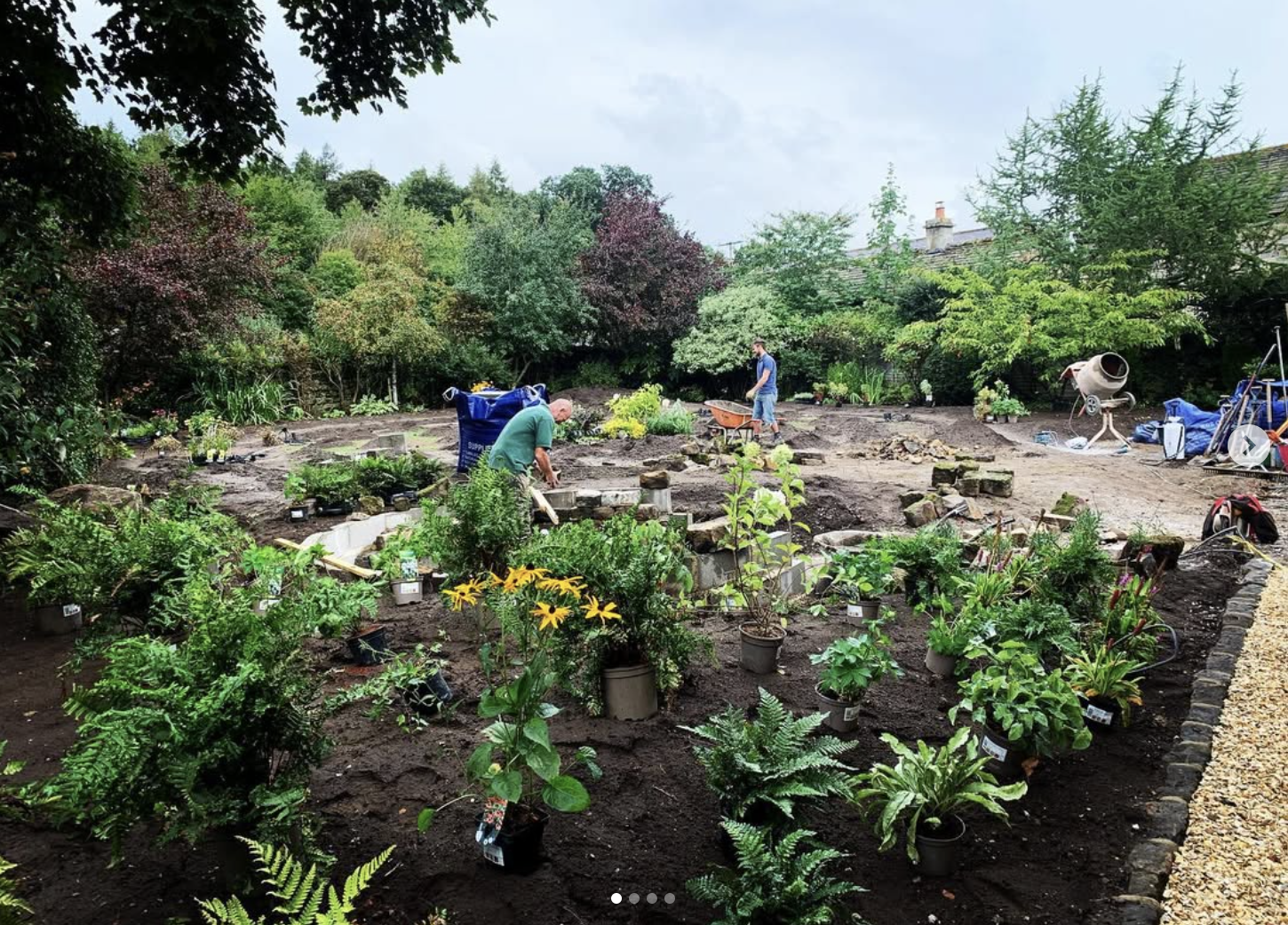 Two men working in a garden under a cloudy sky, planting and arranging plants, with trees and shrubs in the background and construction tools and materials around.