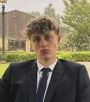 Young man with curly hair wearing a suit and tie outdoors with greenery and a building in the background.
