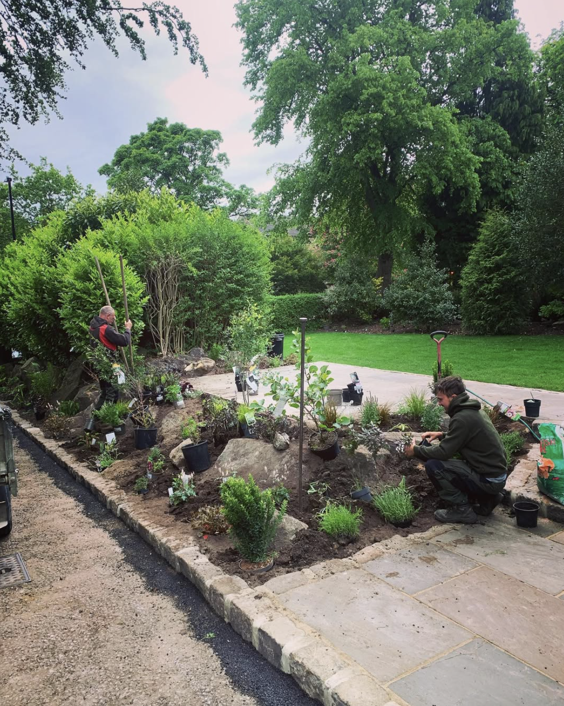 Two people gardening in a backyard with lush green trees and bushes. One person is standing using a long tool, and the other is crouching planting or tending to a shrub. There are gardening tools and potted plants around them.