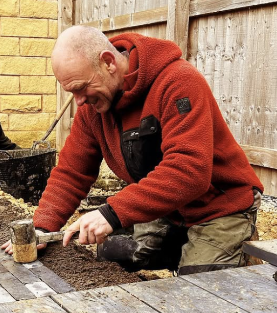 A man in a red fleece jacket and khaki pants is kneeling and working with soil in a garden bed, smiling and using a gardening tool.