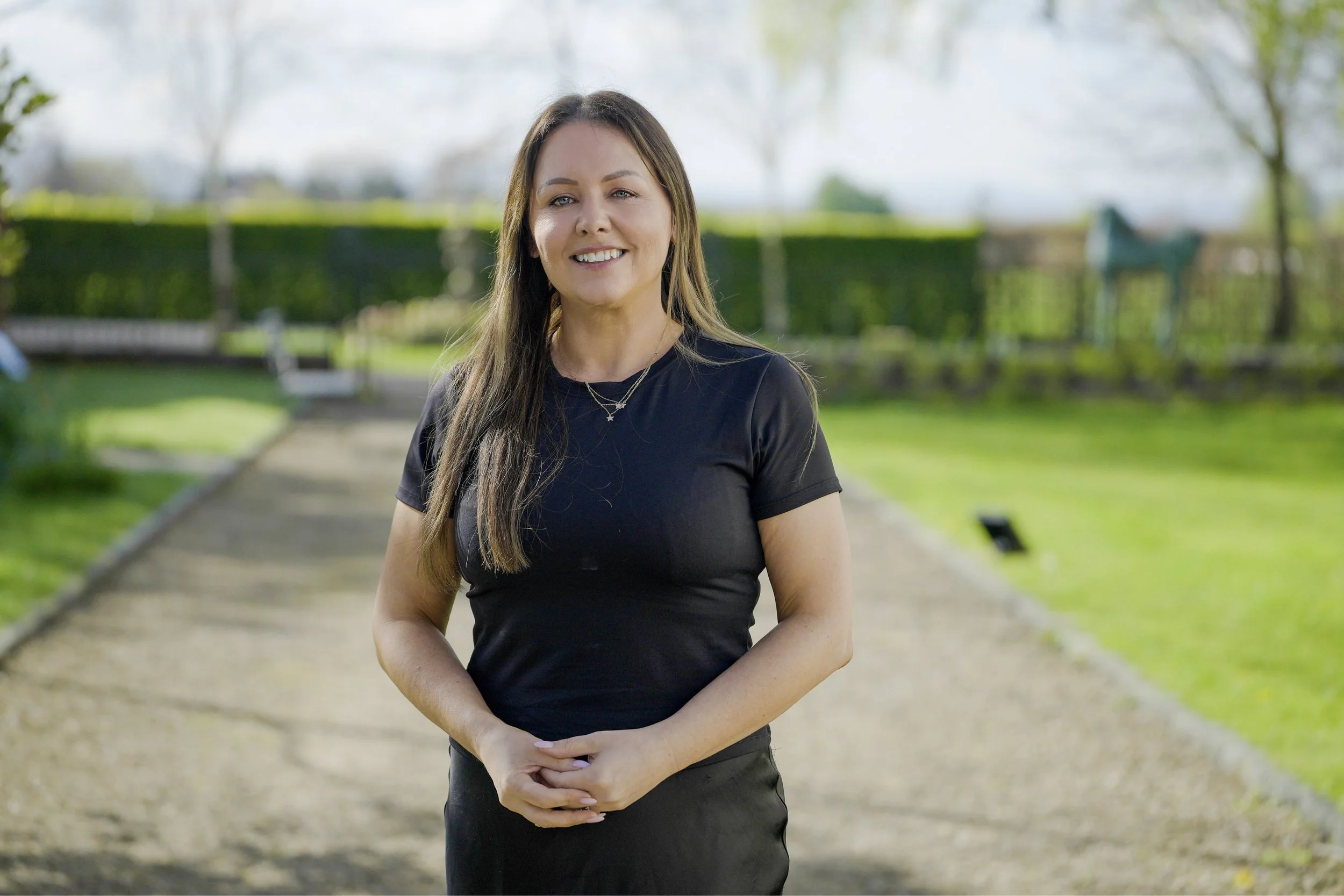 A smiling woman with long brown hair and light makeup, wearing a black t-shirt and layered necklaces, standing outdoors on a pathway in a park with greenery and trees in the background.