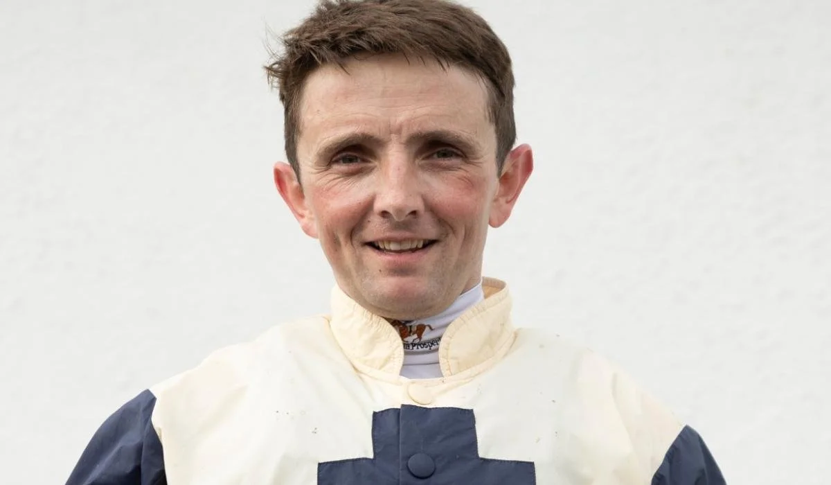A man wearing a racing uniform with a cross on the chest, smiling, with a white background.