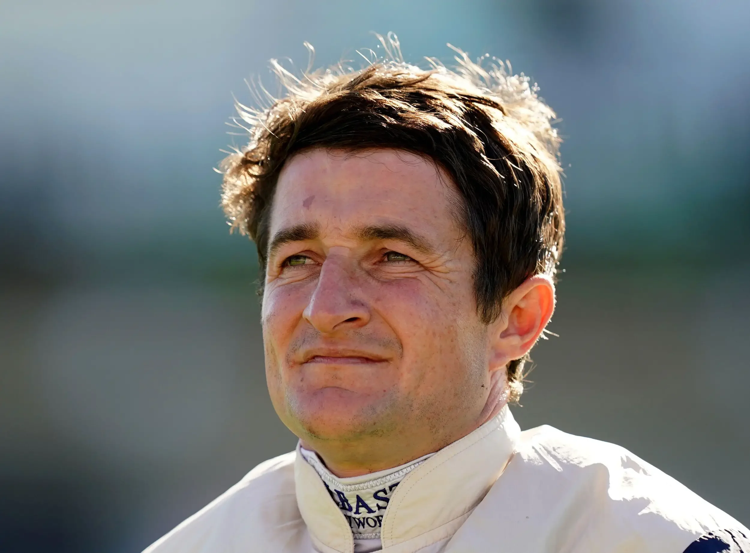 Close-up of a smiling man with short dark hair, wearing a white racing suit, outdoors with a blurred background.