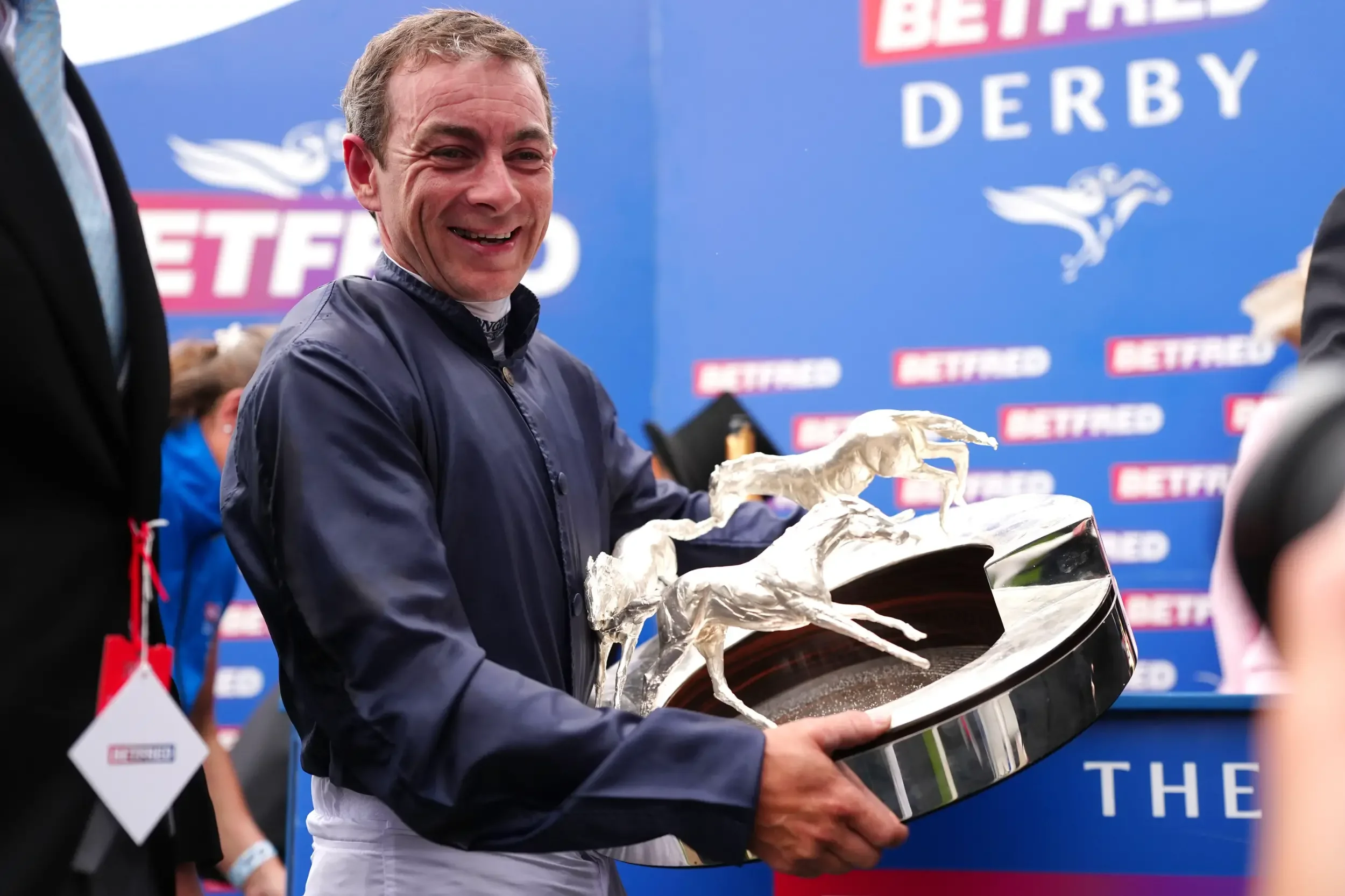 A man holding a trophy shaped like a shoe, smiling at an event with a blue backdrop that reads 'BETFRED DERBY.'