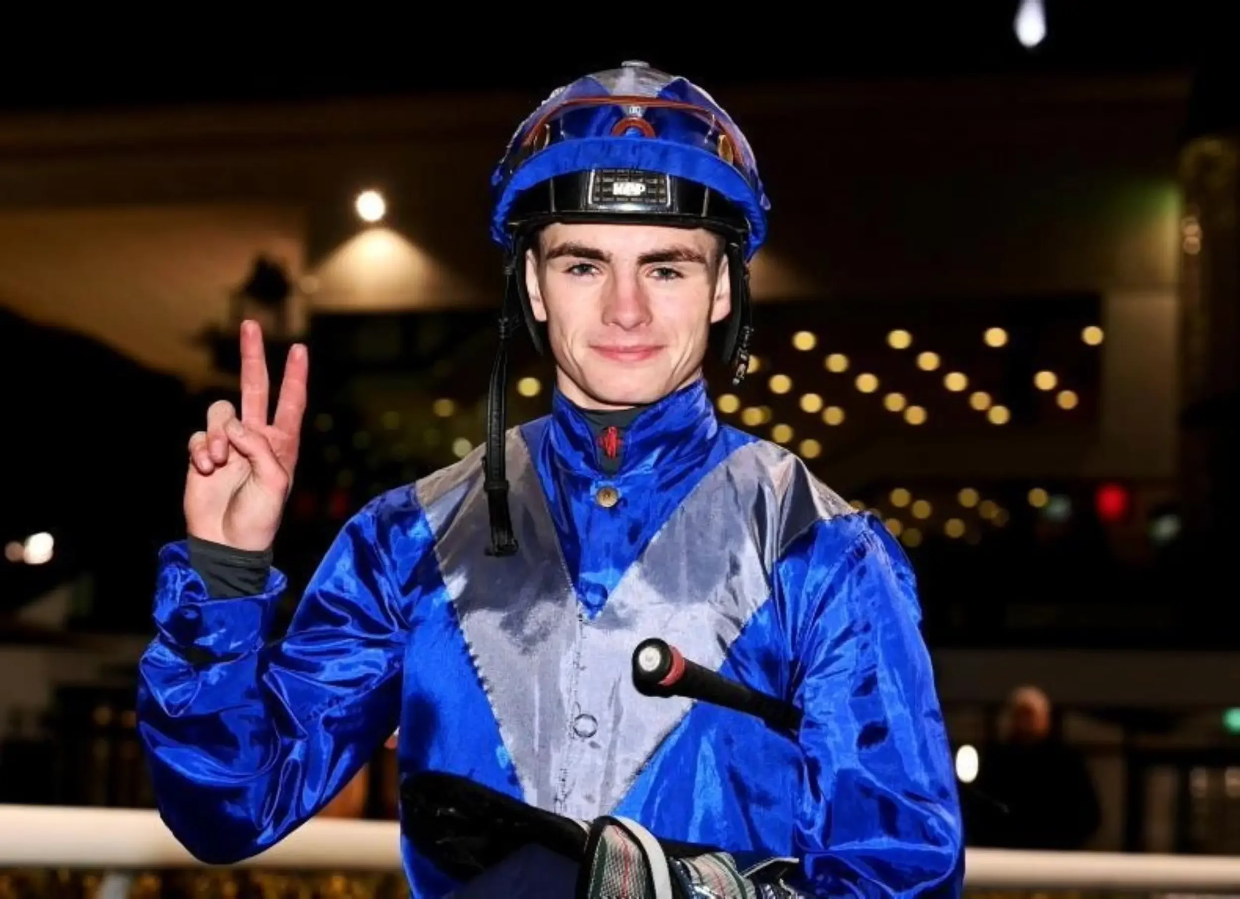 A young man wearing a blue helmet and a blue and gray jacket, smiling and making a peace sign with his right hand, riding a bicycle indoors.