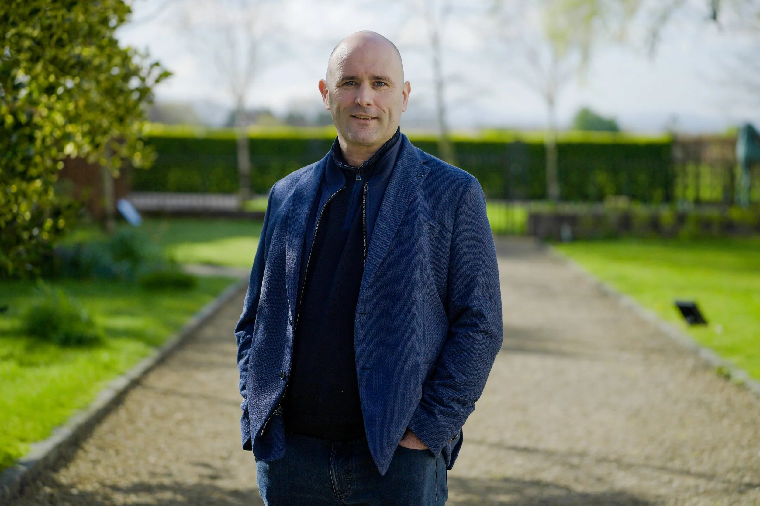 A bald man wearing a blue blazer over a dark zip-up top, standing on a park pathway with green trees and grass in the background on a bright day.