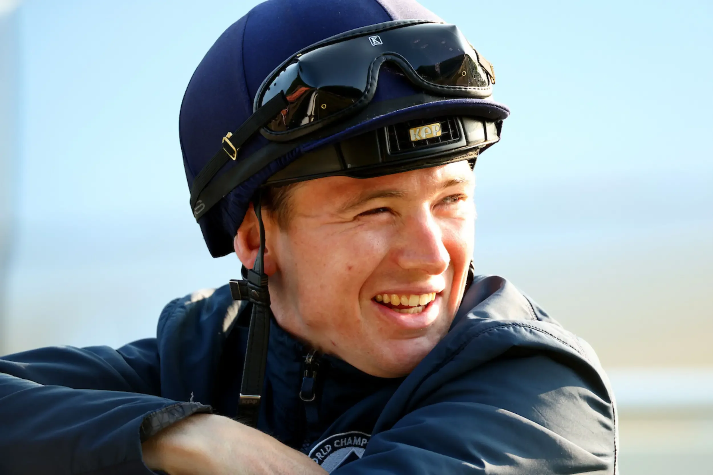 A young man smiling wearing a motorcycle helmet with goggles, dressed in a dark jacket, outdoors with a clear sky.