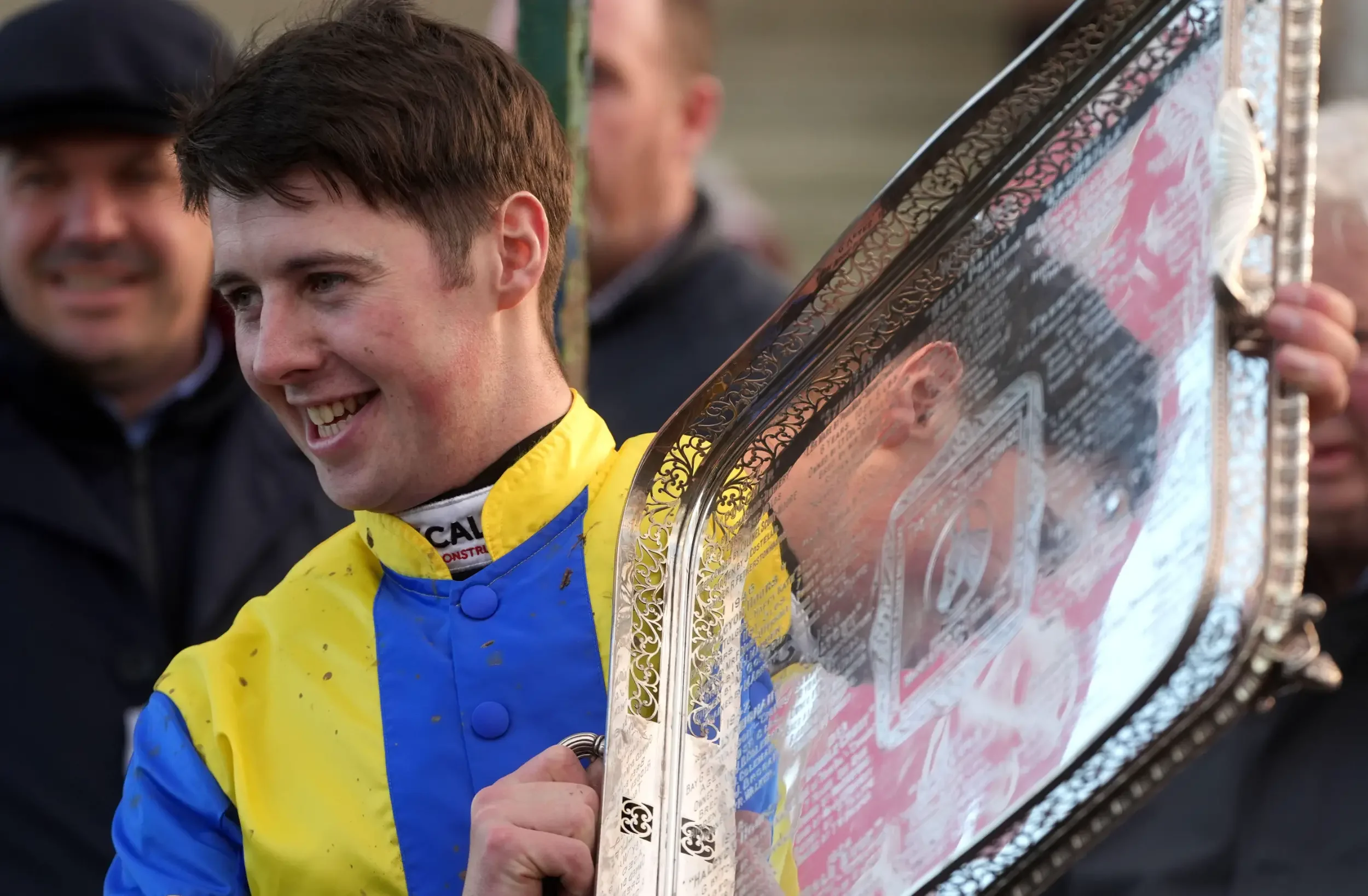 A young male cyclist in a yellow and blue jersey holding a large, decorative trophy with silver and gold details.