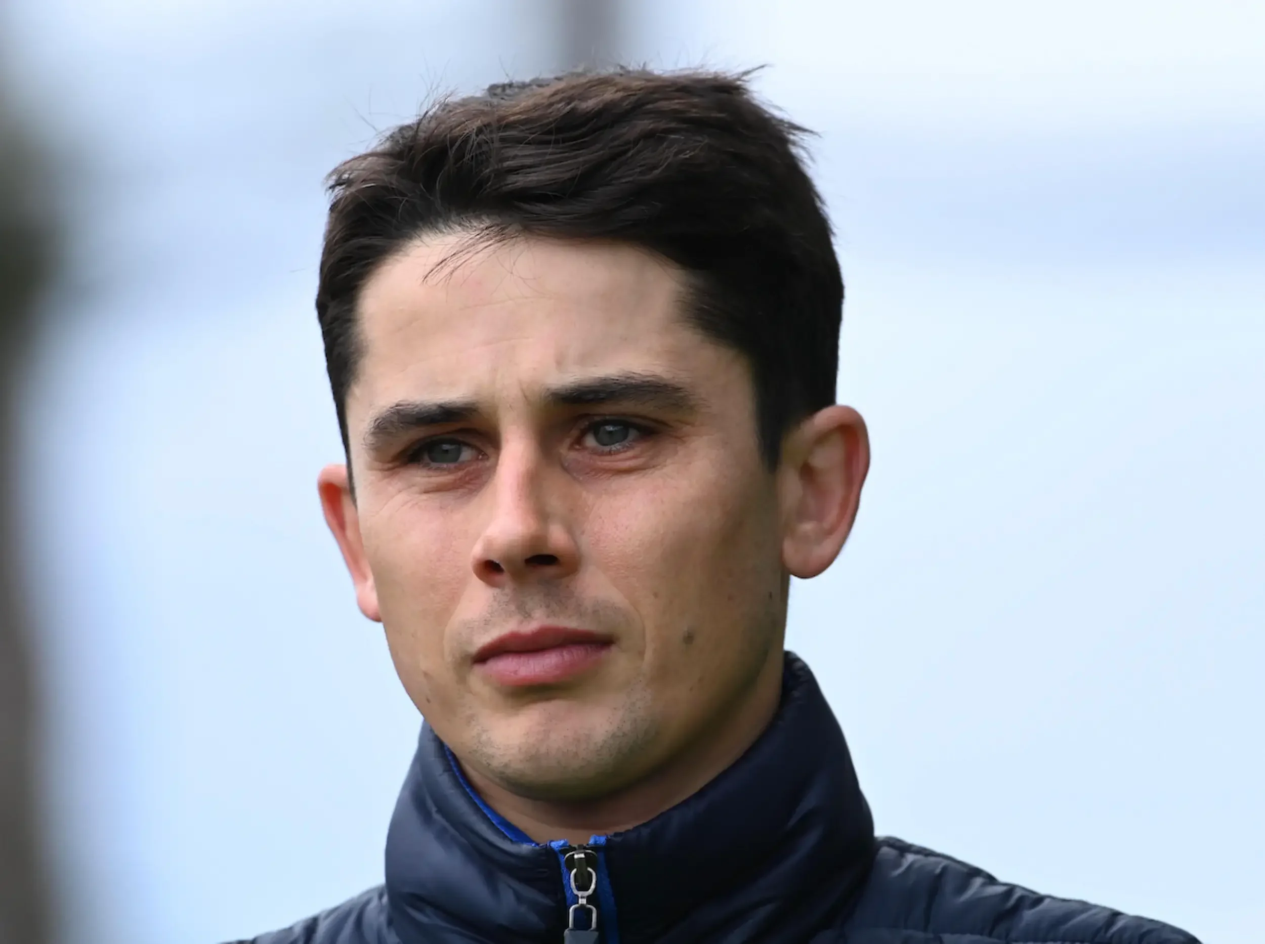 Close-up of a young man with dark hair, blue eyes, and light skin, wearing a dark jacket with a zipper, outdoors against a blurred light blue sky background.