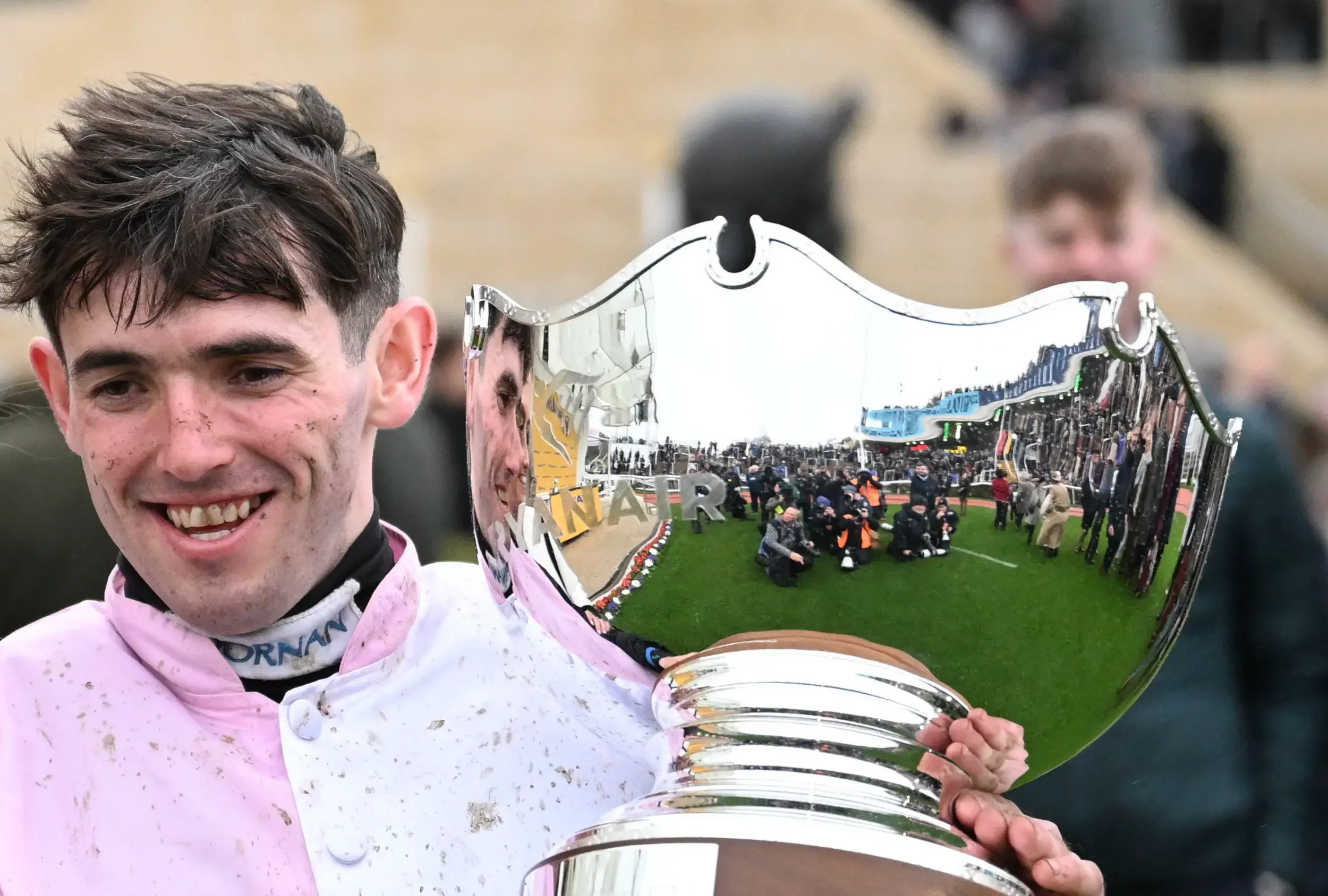 A young man with short dark hair holding a shiny silver trophy, wearing a pink and white racing suit with mud stains. He is smiling and standing outdoors at a race track, with spectators and photographers in the background.