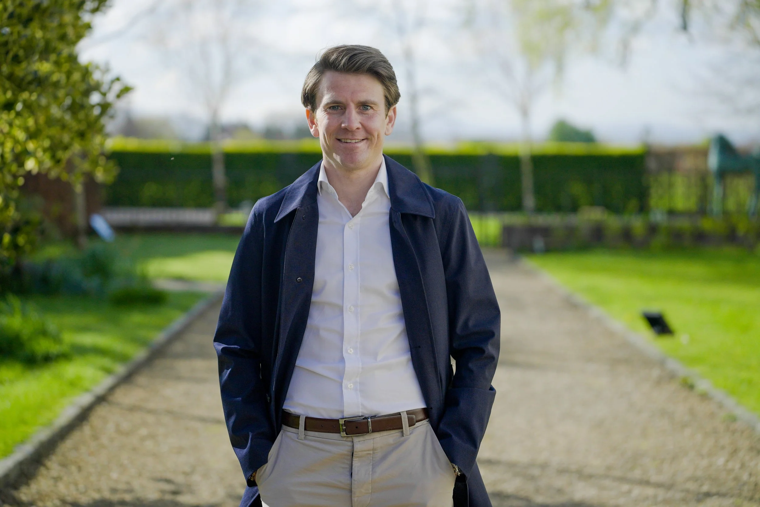 A man with brown hair, blue eyes, wearing a white shirt, dark jacket, and beige trousers, standing on a garden pathway with green trees and a hedge in the background.