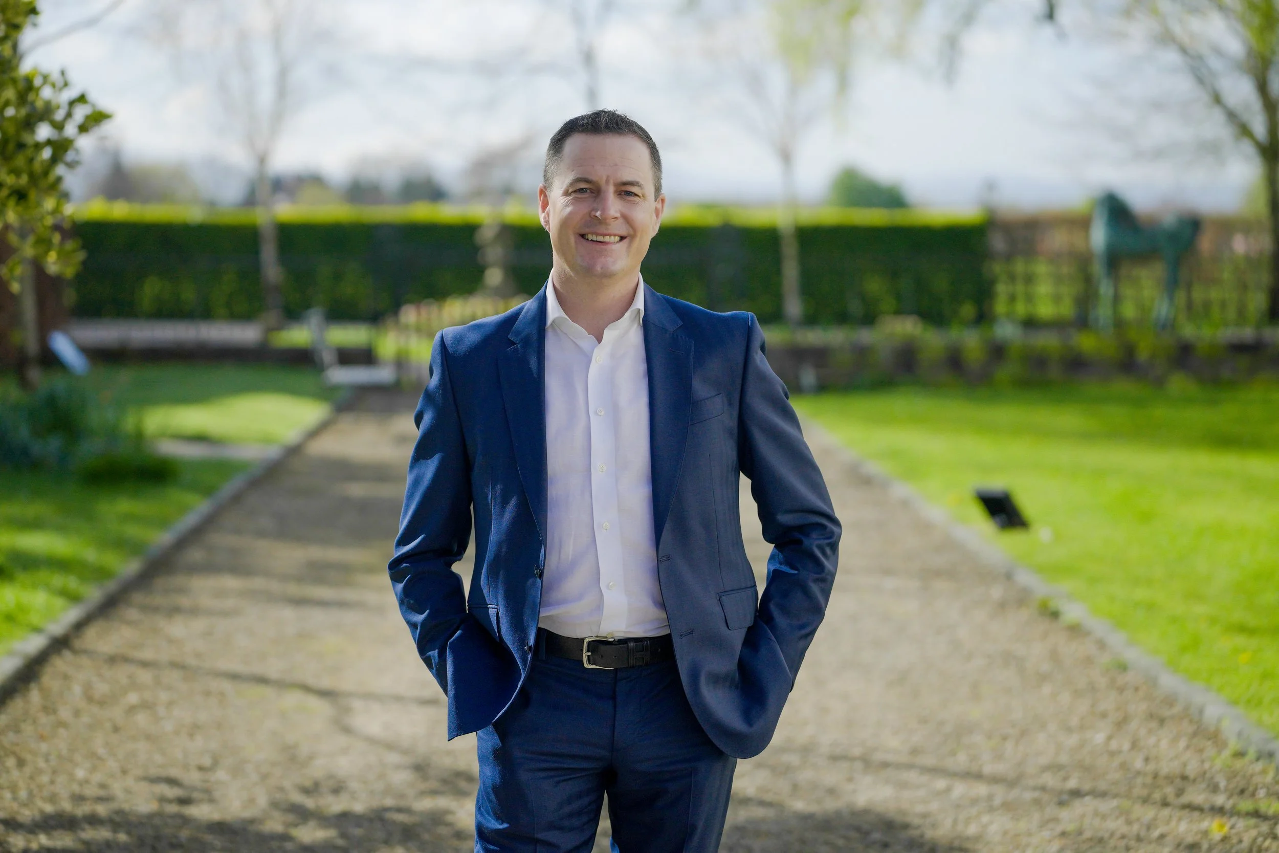 A man in a blue suit and white shirt is smiling and standing with his hands in his pockets on a park pathway during daytime with trees and greenery in the background.