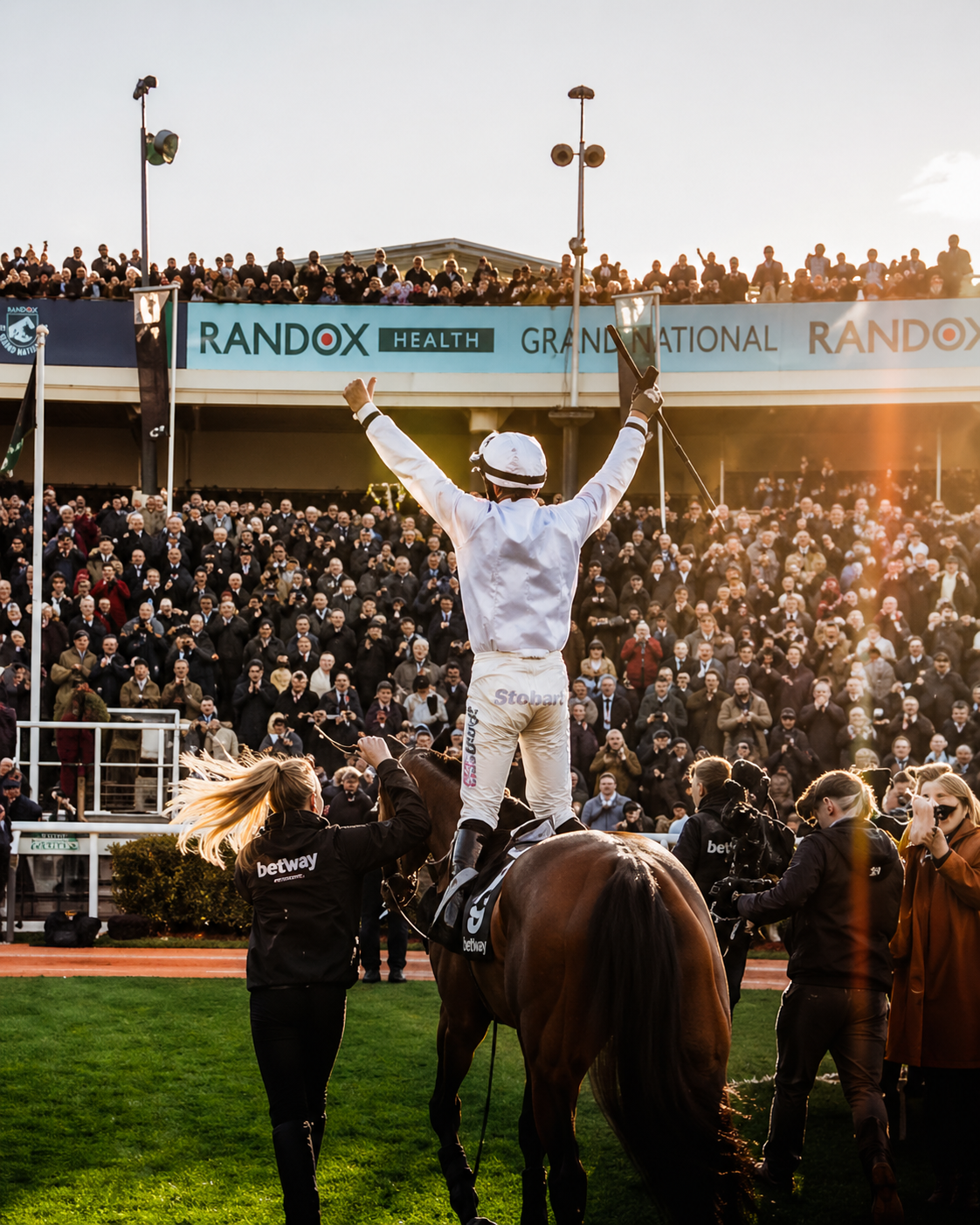 A jockey standing on a rearing horse, with arms raised in victory, surrounded by a crowd at a racetrack.