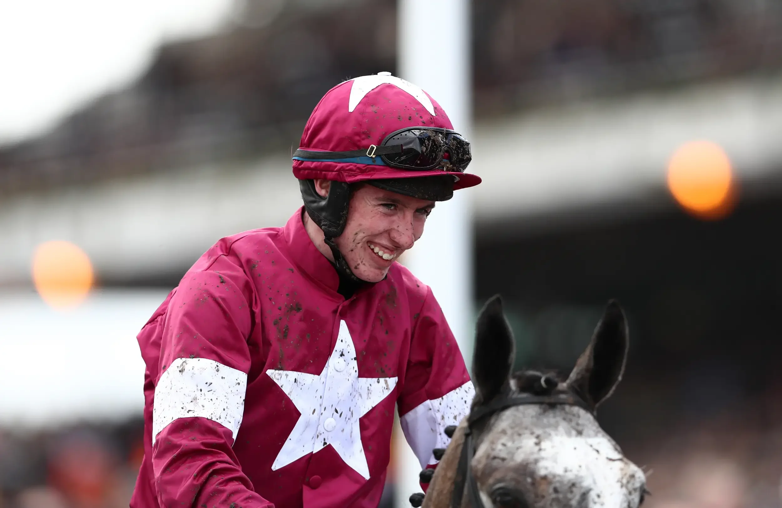 A young male jockey with a pink and white silks riding outfit and black goggles on his helmet smiling while holding a white and brown speckled horse.
