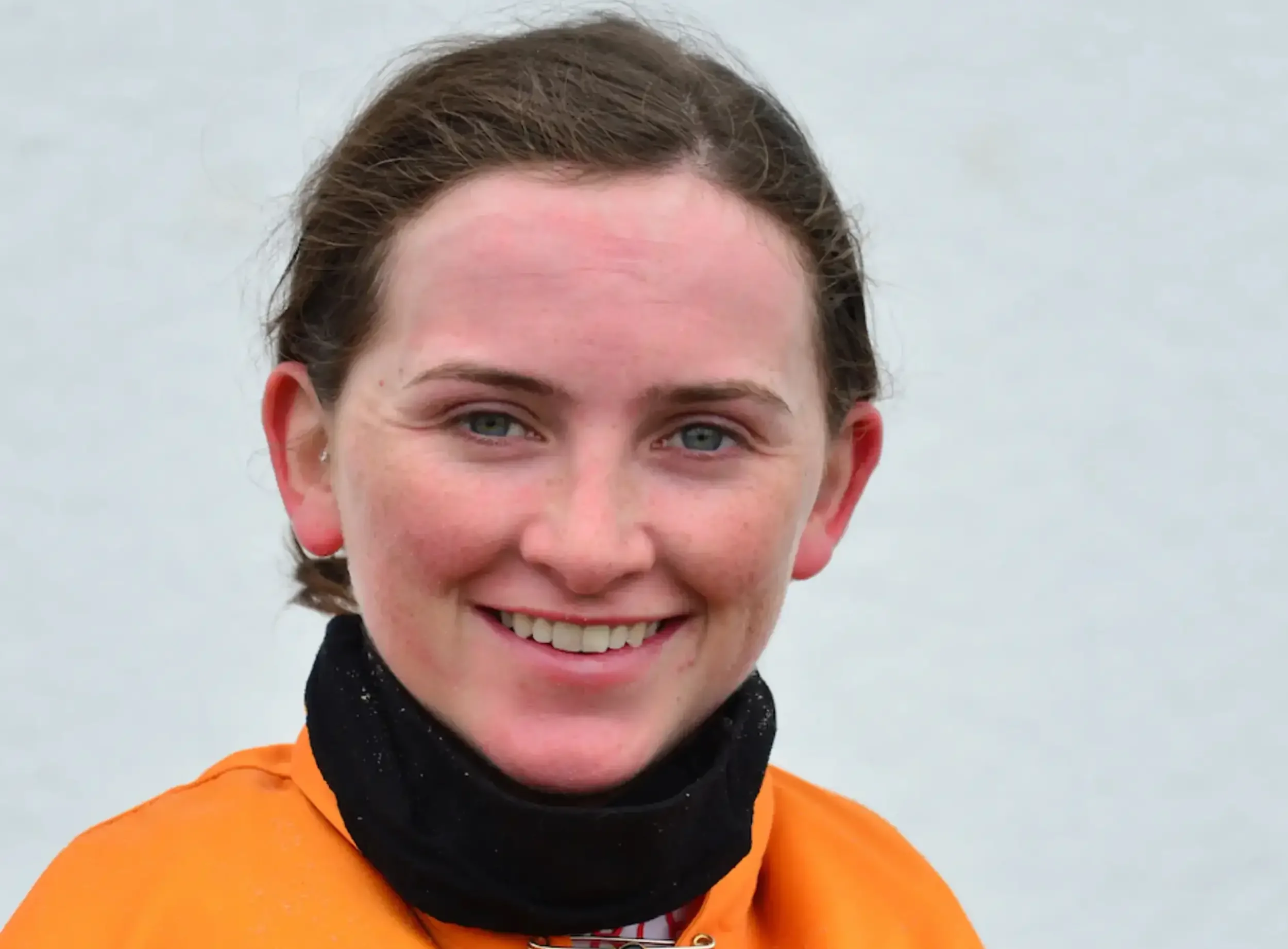 Close-up of a smiling woman with blue eyes and brown hair in a bun, wearing an orange jacket and a black neck gaiter, against a plain light background.