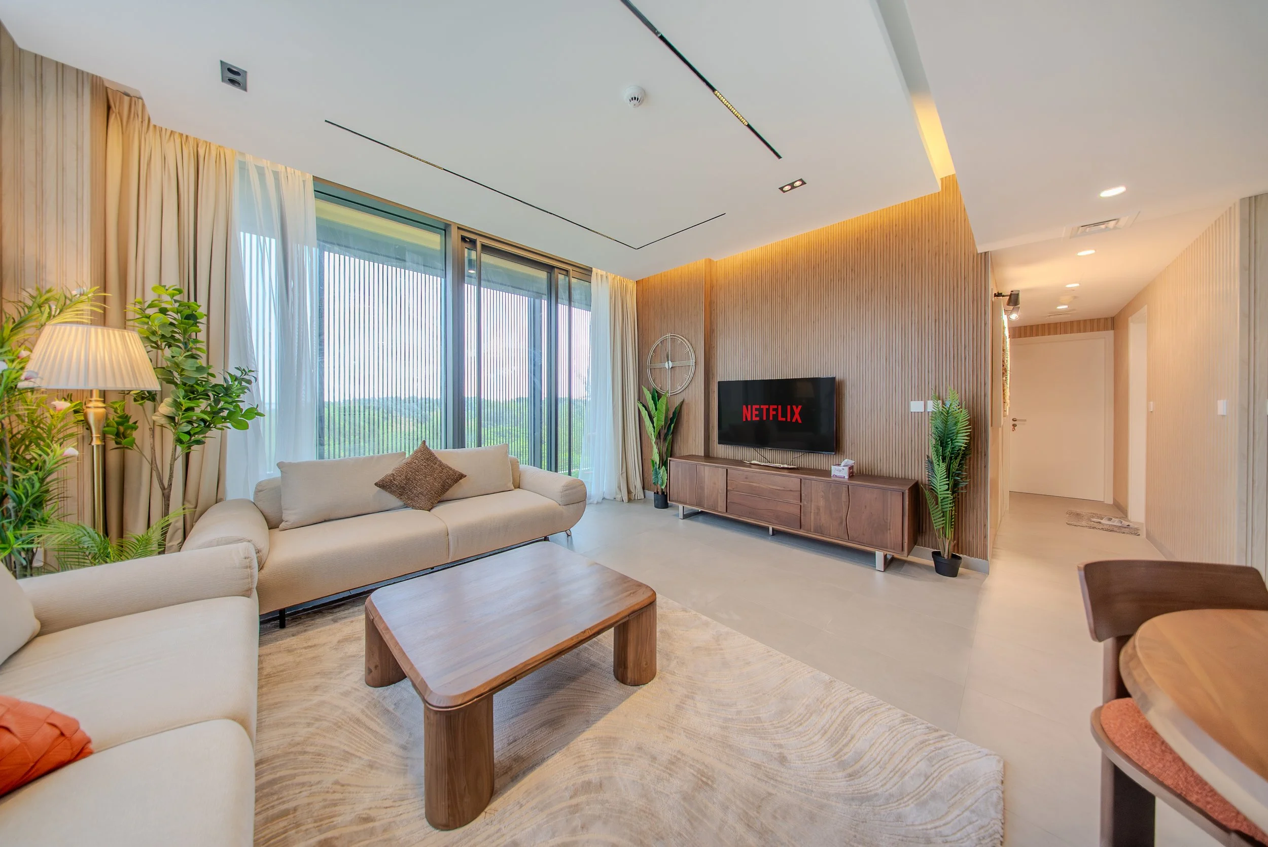 A modern living room with white sofas, a wooden coffee table, potted plants, a flat-screen TV displaying 'Netflix,' large windows with sheer curtains, and wood-paneled walls with warm lighting.