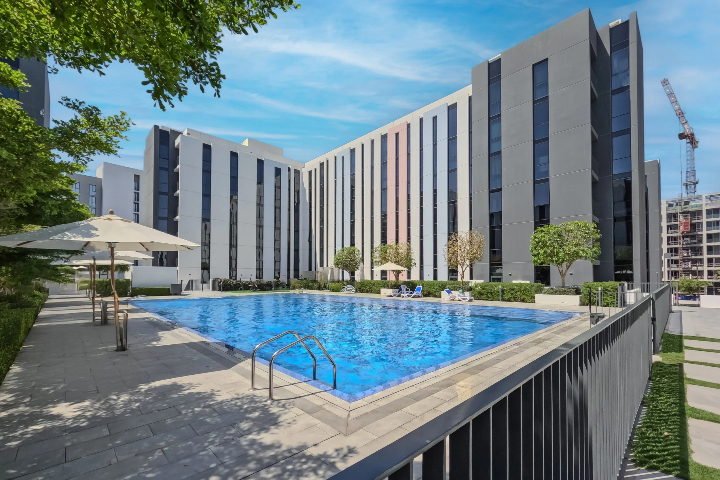 Modern apartment complex with a swimming pool in the foreground, surrounded by trees and shaded lounge chairs, with a construction crane visible in the background.