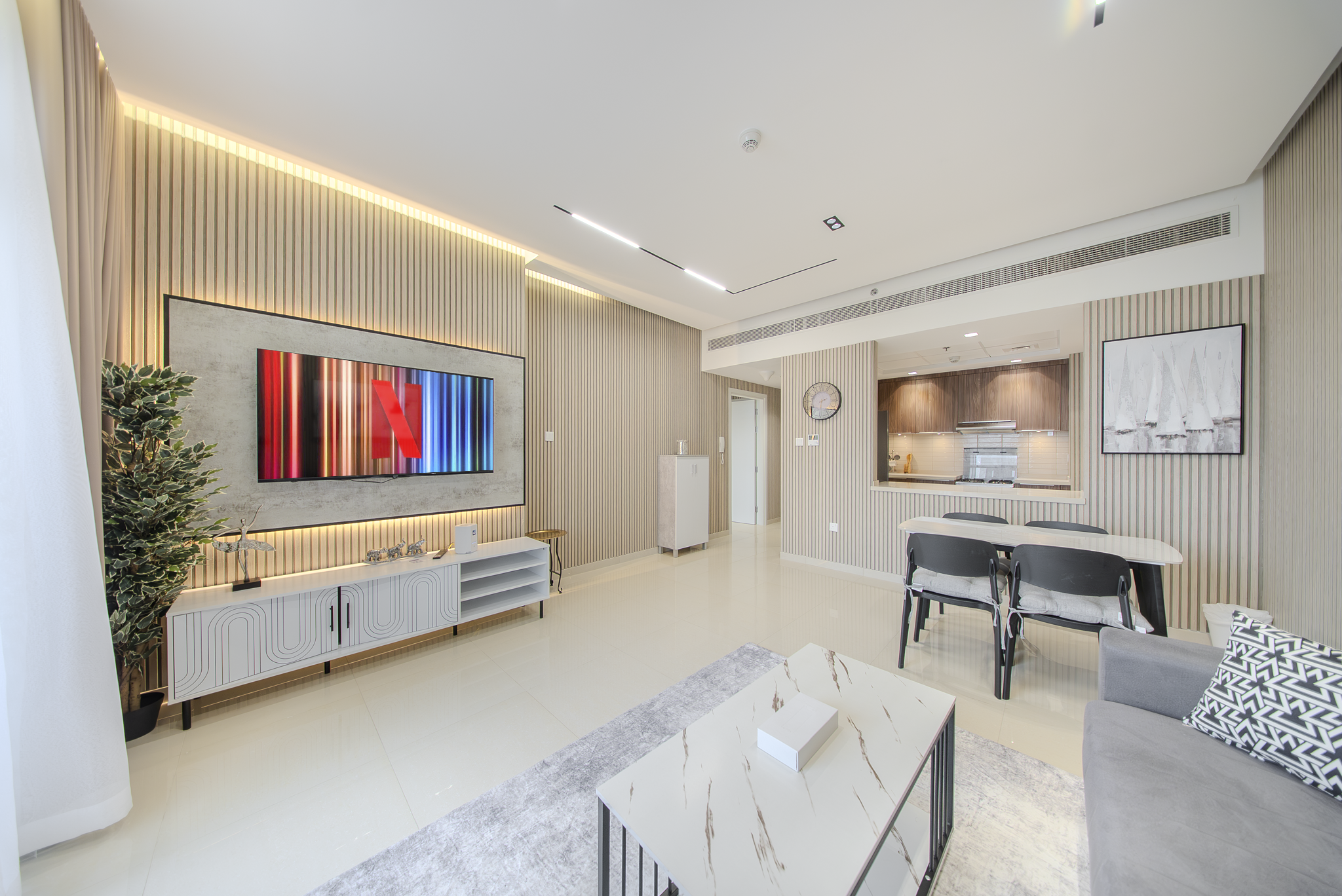 Modern living room with beige striped walls, a white TV console, a large flat-screen TV, a grey couch with patterned cushions, a marble coffee table, and a dining area with black chairs and a white table. The room has a minimalist design with bright, natural lighting.
