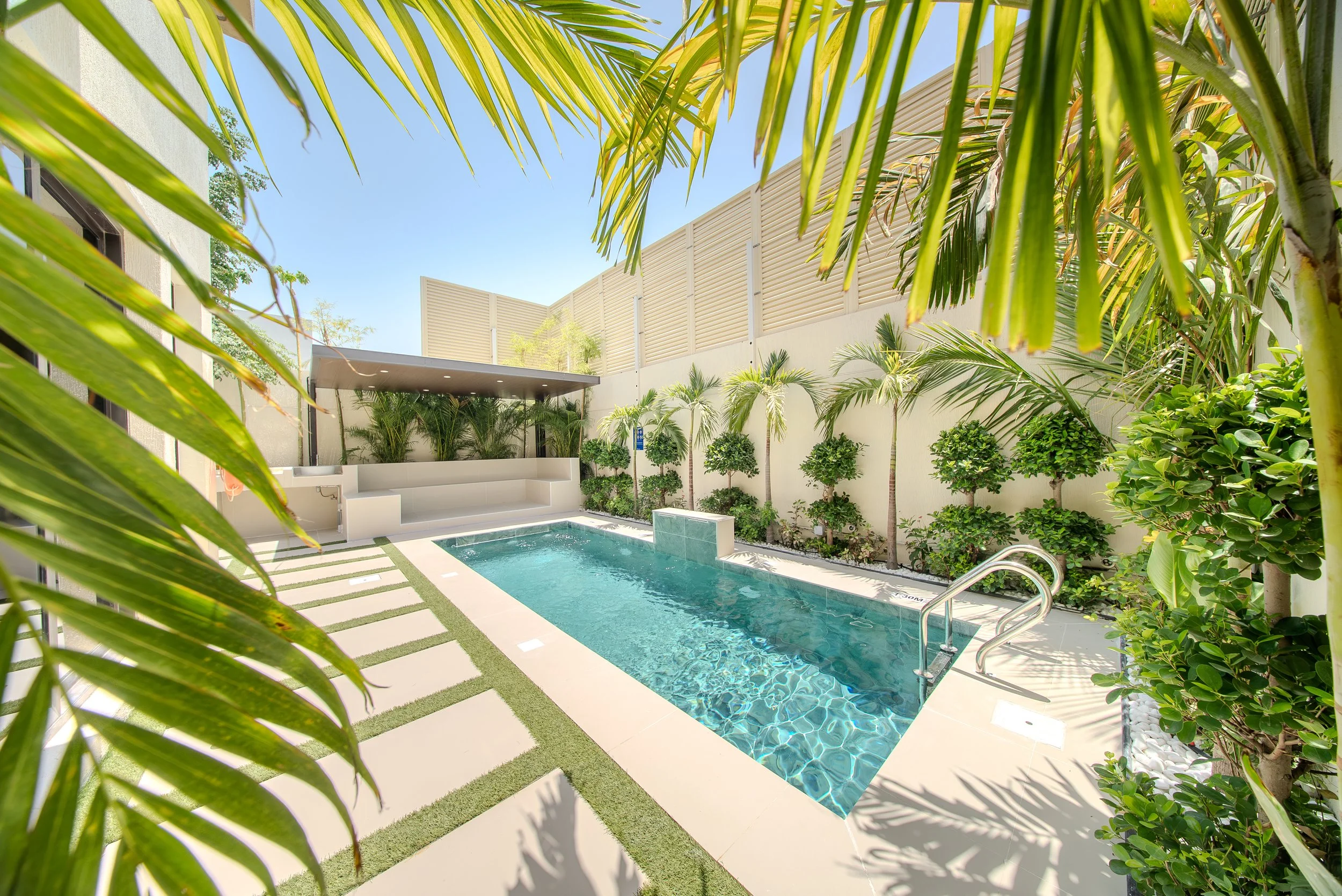 A backyard with a private swimming pool surrounded by tropical plants and trees, with a high white fence and beige building walls in the background, under a clear blue sky.