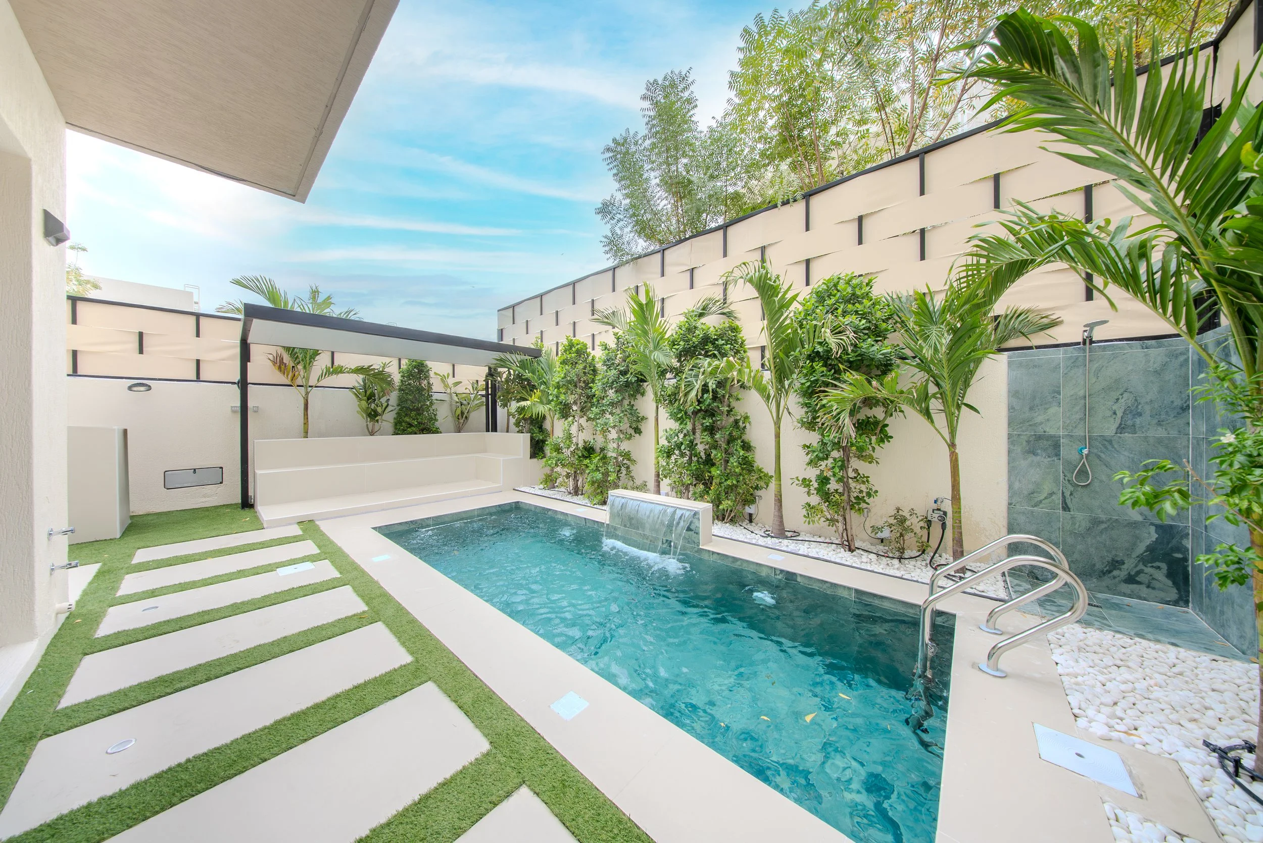Modern backyard with a small rectangular swimming pool, surrounded by a white patio, lush greenery including various plants and small trees, and a high cream-colored fence. The sky is partly cloudy.