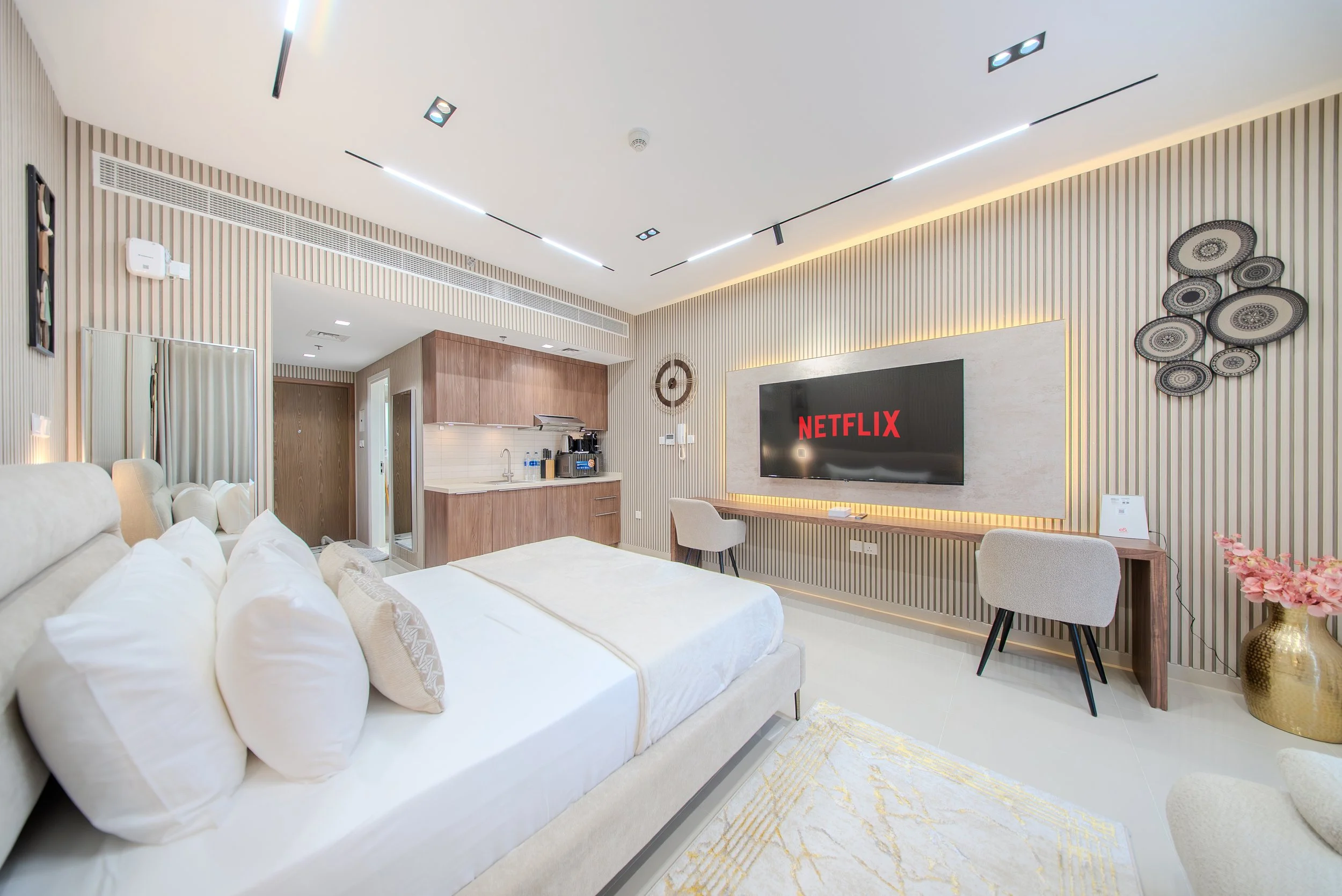 Modern living room with a large bed, white bedding, beige chairs, a wall-mounted flat-screen TV displaying NETFLIX, decorative artwork, a kitchenette, and gold accents.