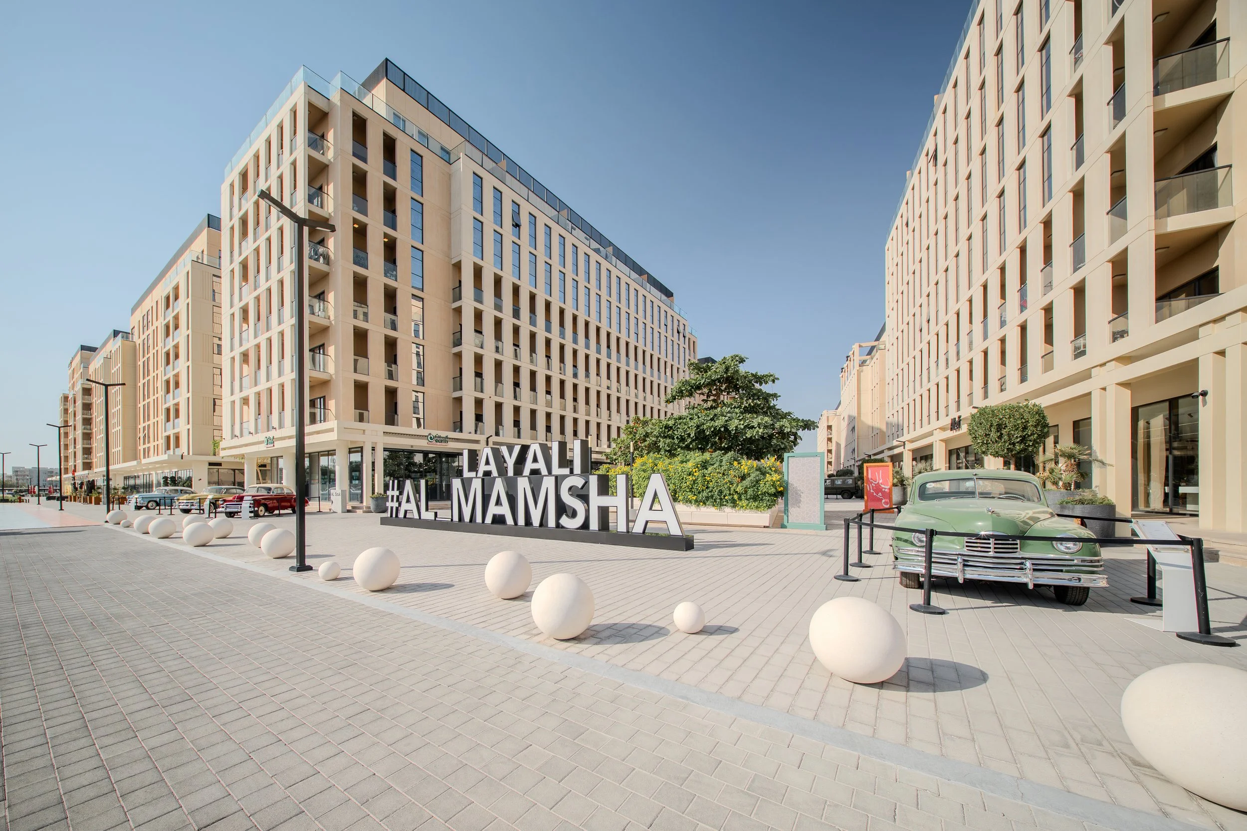 Modern beige apartment buildings with glass balconies under a clear blue sky, a vintage green car parked along the sidewalk, and a plaza with large white sphere decorations and a sign reading '#HALMAMSHA'.