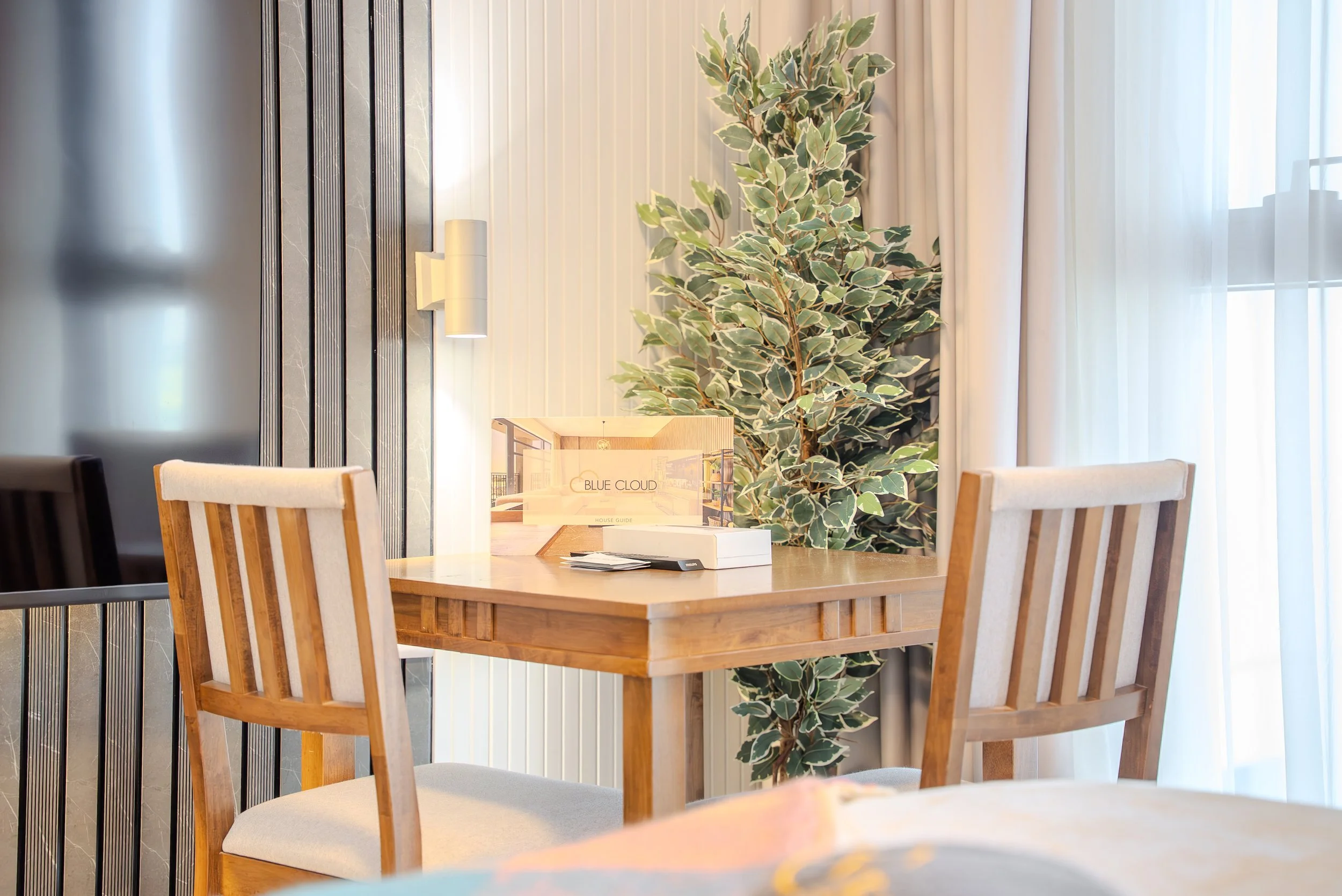 A dining area with a wooden table and four wooden chairs, a large green houseplant, and curtains covering a window, with a softly-lit wall decor and a house guide brochure on the table.