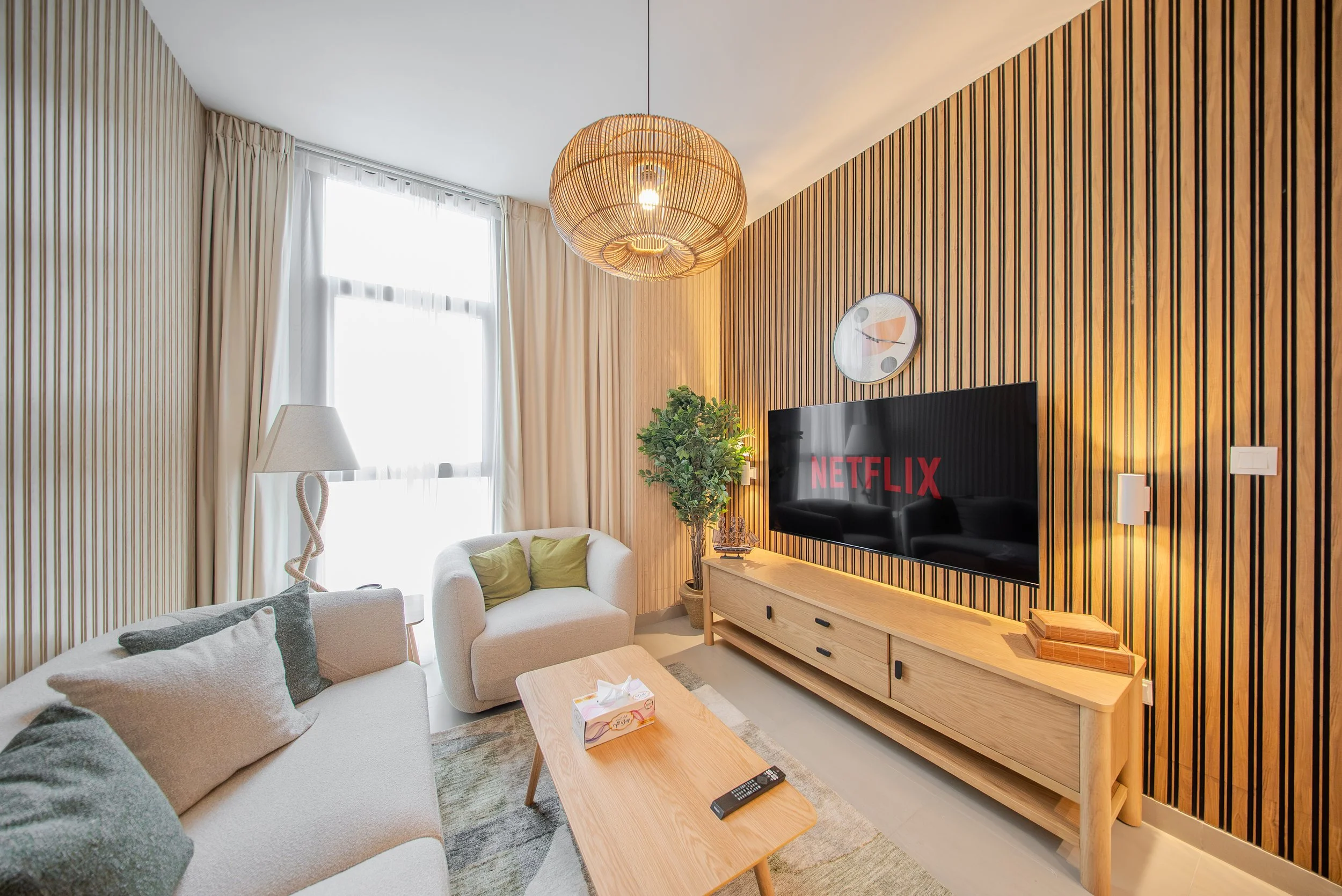 Living room with cream sofas, beige and green pillows, wooden coffee table, potted plant, TV with Netflix logo, wood slatted accent wall, round wall clock, large window with white curtains, modern hanging light fixture.