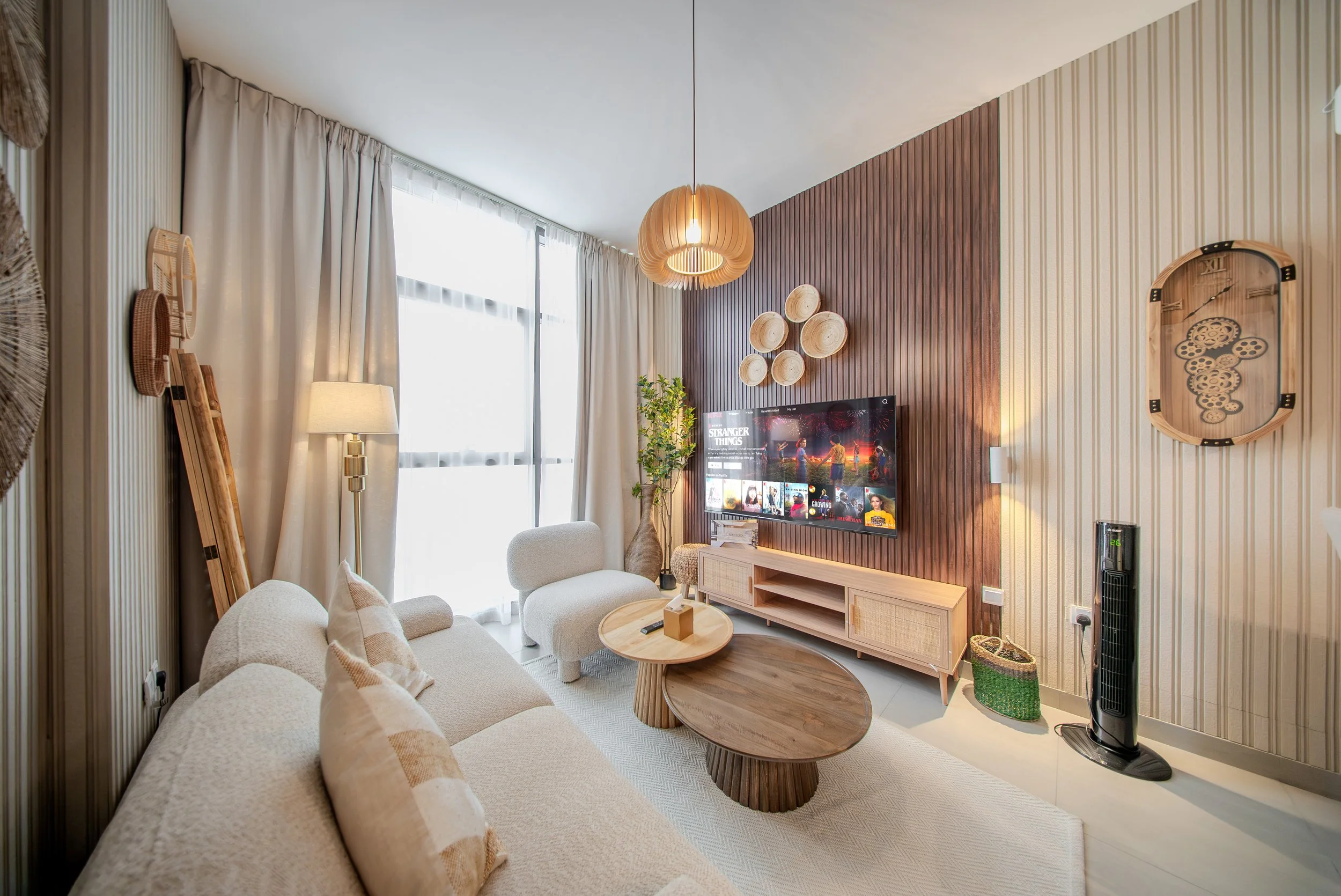 Living room with beige sofa, wooden coffee table, wall-mounted TV, wooden wall panel, decorative wall art, standing fan, and floor lamp near window with white curtains.
