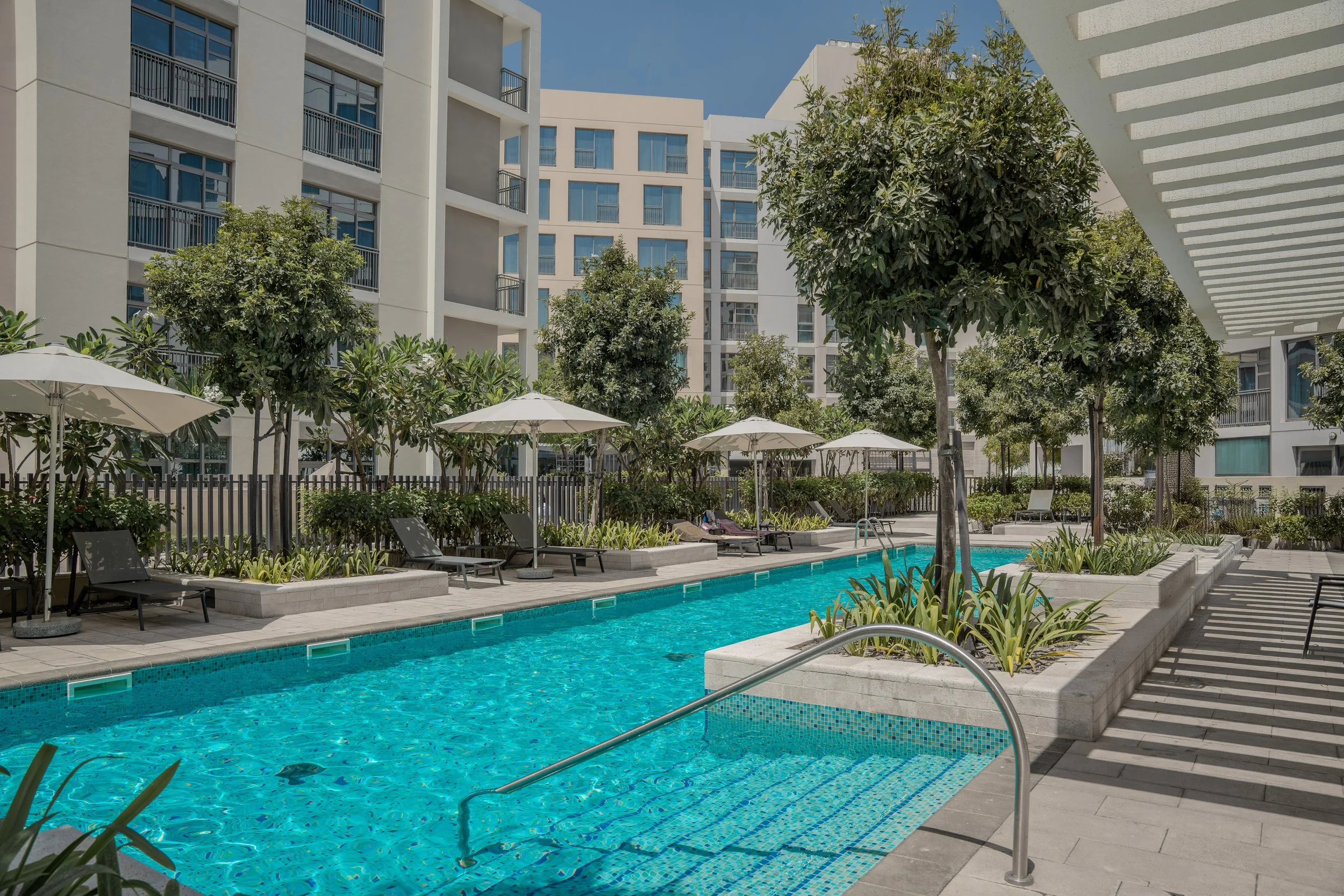 A swimming pool surrounded by lounge chairs, umbrellas, and trees in a modern apartment complex courtyard.