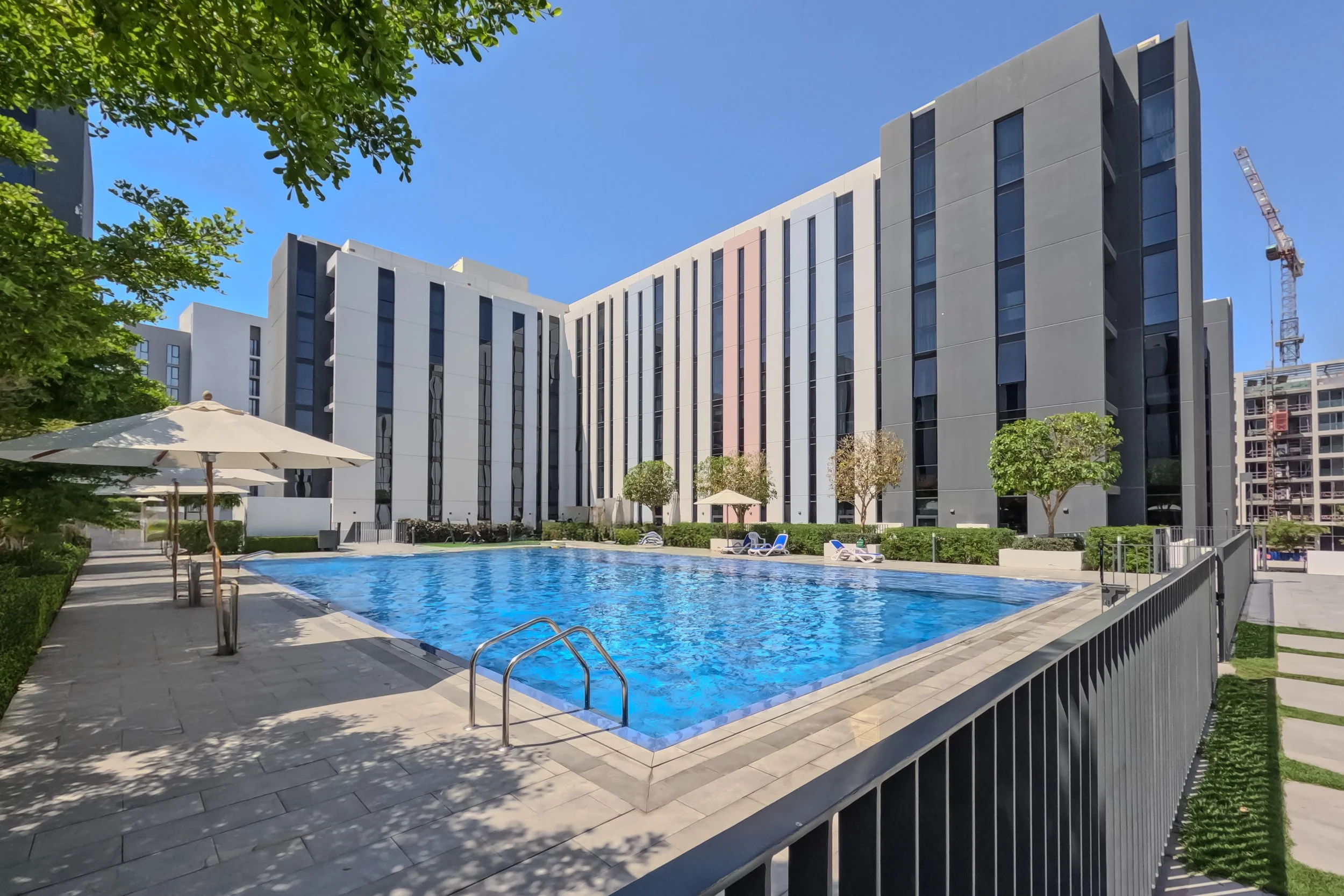 Modern apartment complex with a swimming pool, poolside lounge chairs, umbrellas, trees, and a construction crane in the background under a clear blue sky.