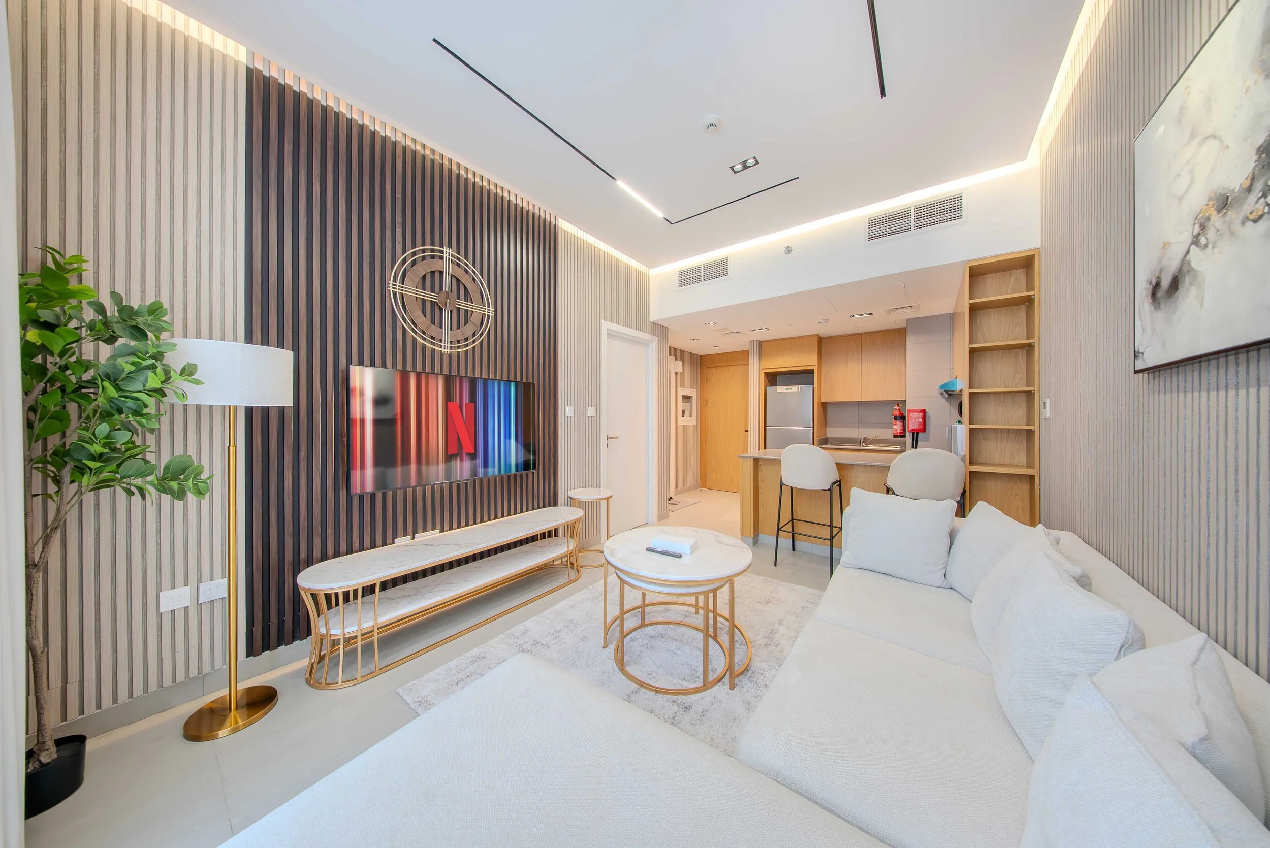 Living room with white sofa, gold and glass coffee tables, TV on a wood-paneled wall, modern clock, green potted plant, and artwork on the wall, with a kitchen area in the background.