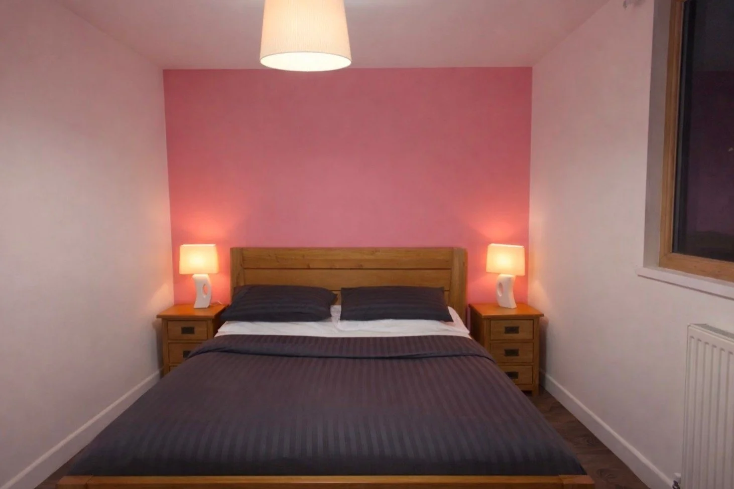 A bedroom with a wooden bed and dark gray bedding, two black pillows, pink accent wall behind the bed, two matching bedside tables with lamps, a ceiling light, and a window on the right side.