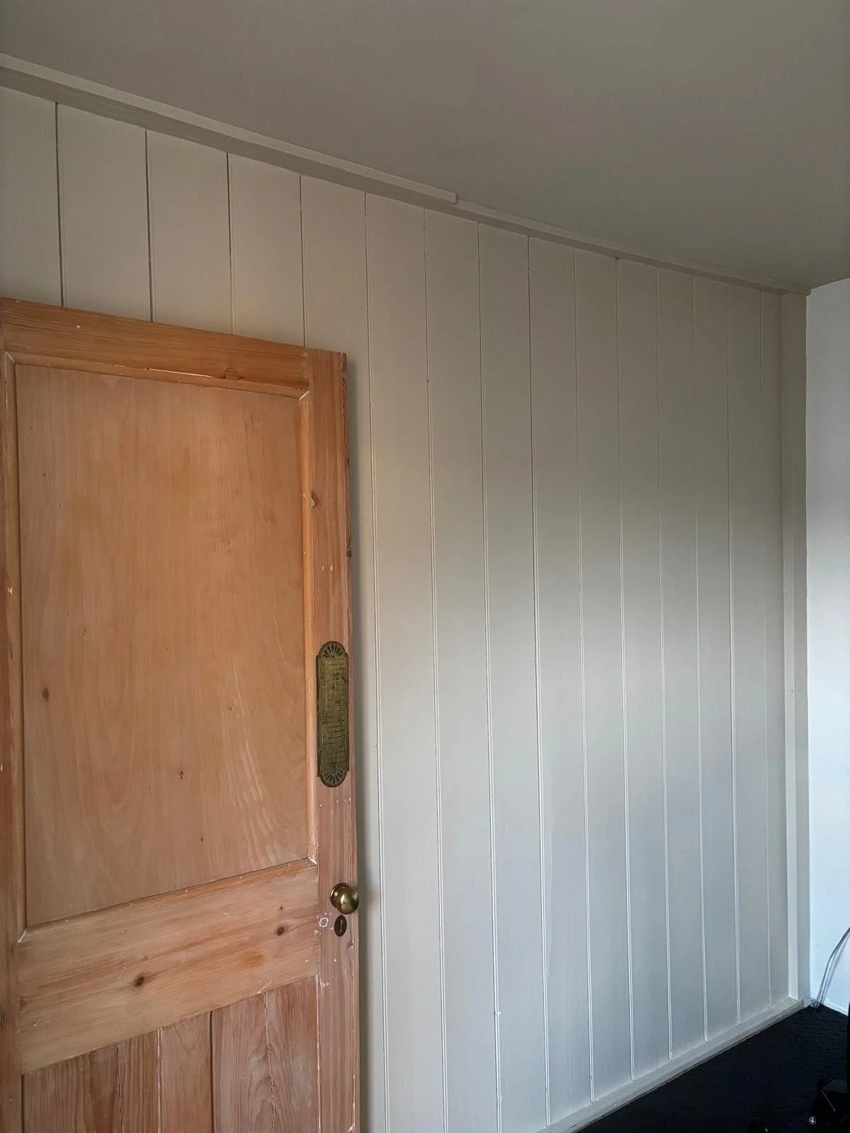 Interior image of a wooden door with a brass doorknob, next to a beige wainscoted wall, with a black carpeted floor and a visible power cord on the right.