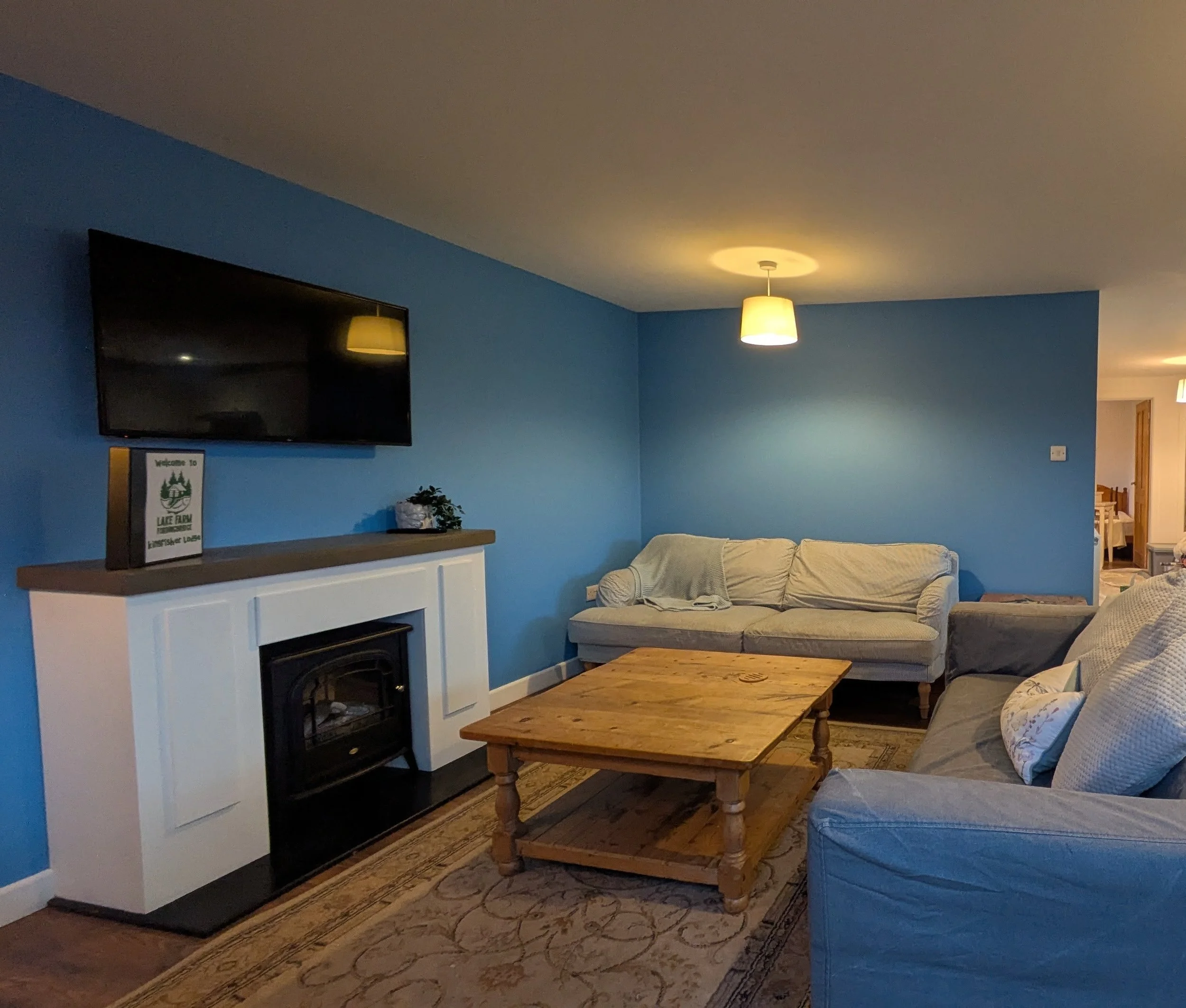 Living room with blue walls, light-colored sofa, wooden coffee table, mounted TV screen, fireplace, ceiling light, and a corner with a sign reading "Welcome to Lake Farm."