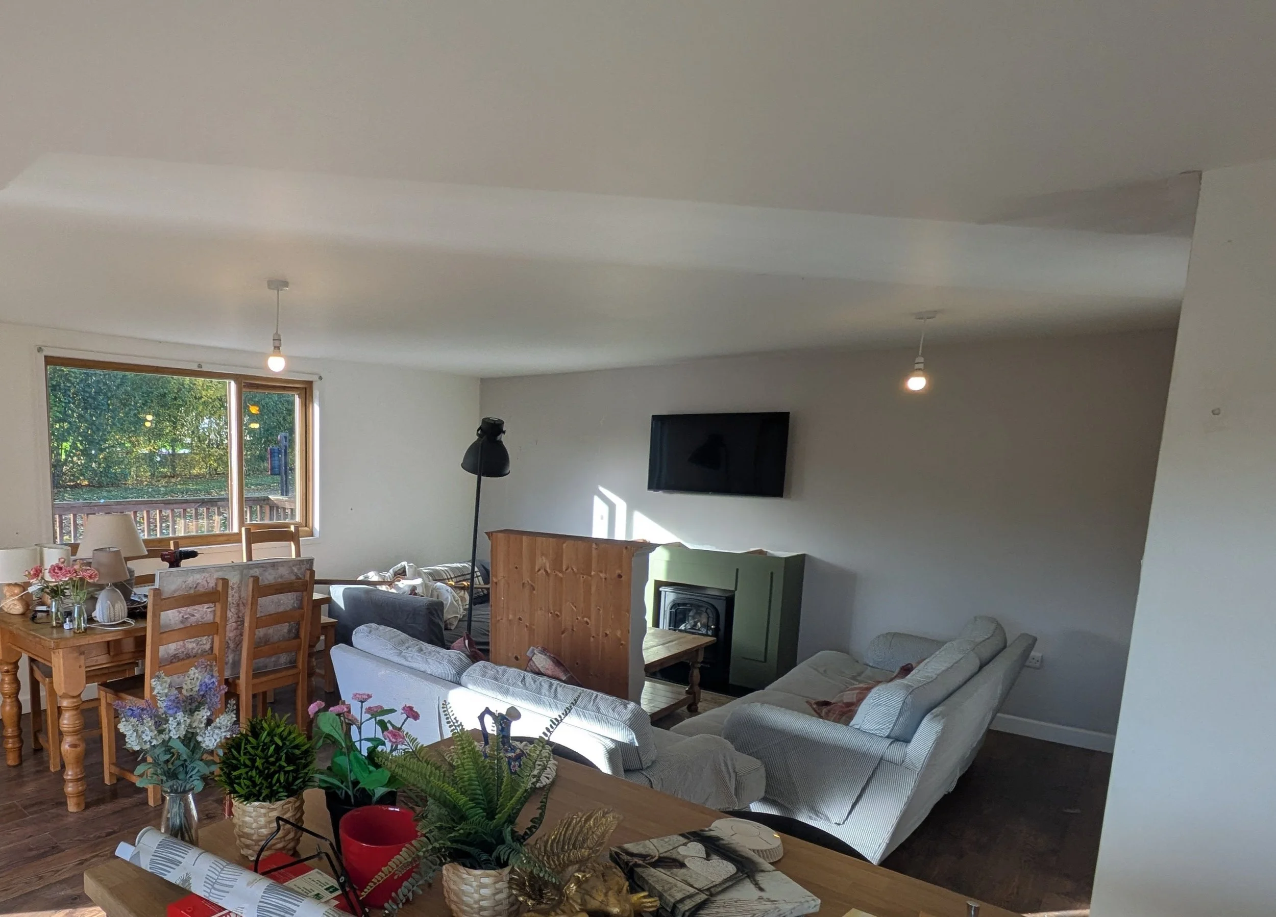 A cozy living room with white walls and large windows showing greenery outside. There are two light bulbs hanging from the ceiling, a wall-mounted TV, a fireplace, and various furniture including sofas and chairs. A dining table with flowers and miscellaneous items is in the foreground.