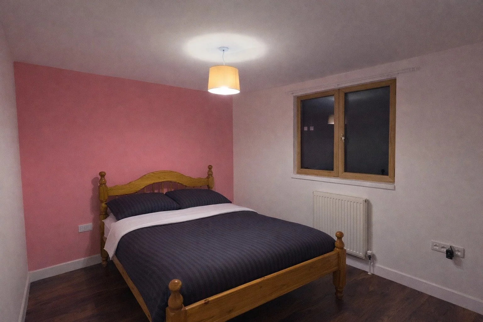 Bedroom with a wooden bed, navy blue striped bedding, two pillows, pink accent wall, window, radiator, and ceiling light.
