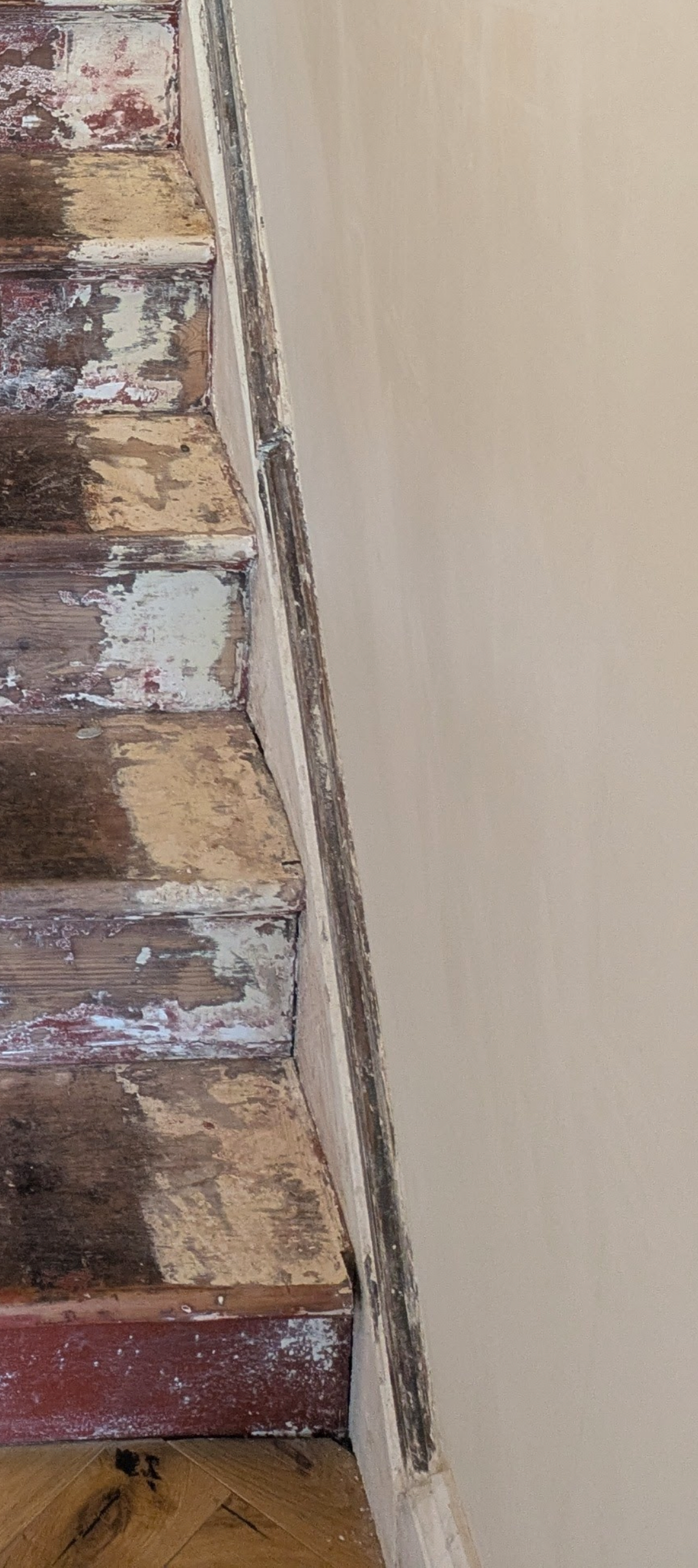 Close-up of a wooden staircase with peeling paint, worn treads, and damaged trim, next to a plain beige wall.