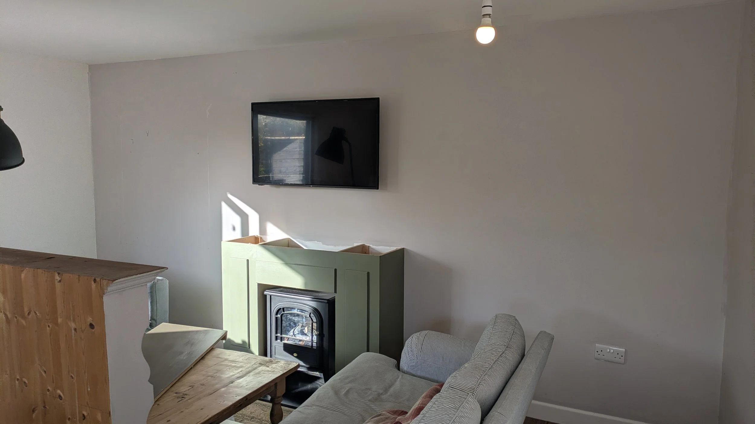 Living room with a wall-mounted flat-screen TV, a small green cabinet, a wood stove, a gray patterned couch, and a wooden dining table, with a ceiling light bulb.