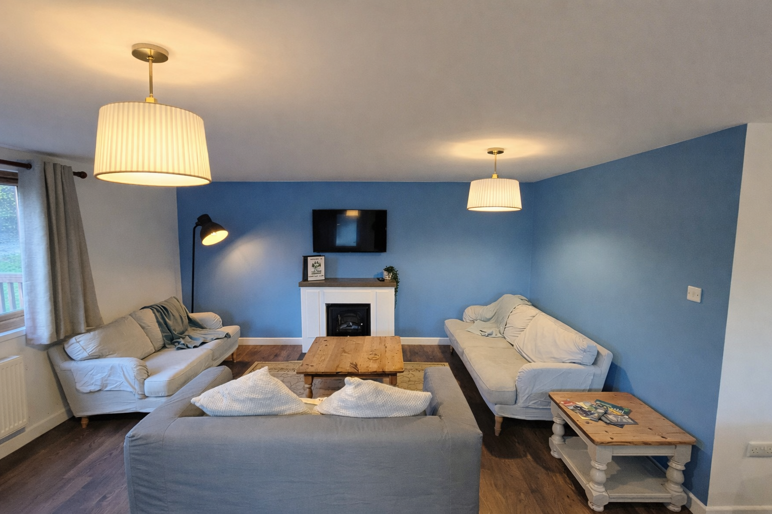 Living room with blue accent wall, beige sofas, wooden coffee table, TV mounted on wall above white fireplace, black floor lamp, window with curtains, and small side table with magazines.