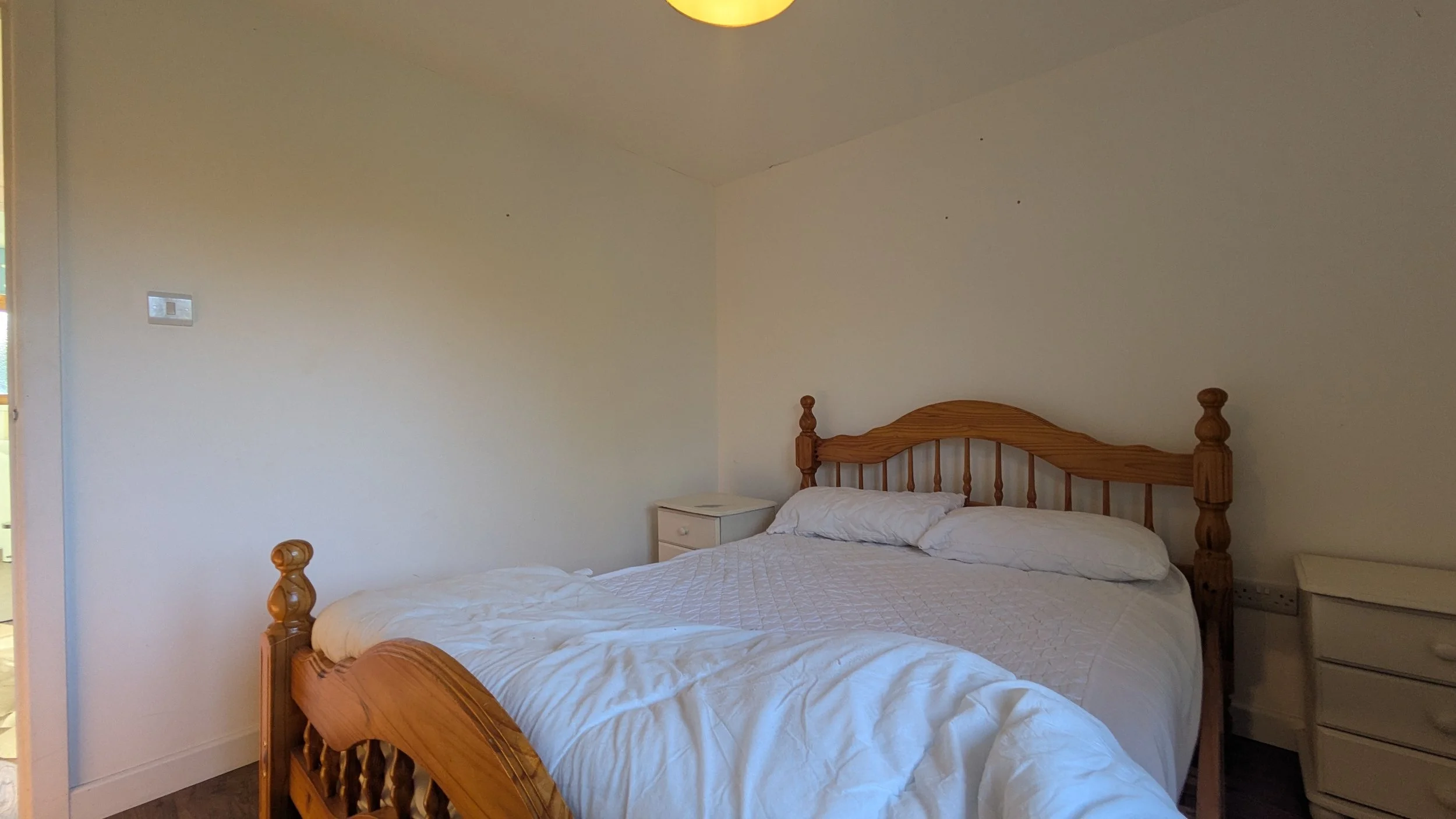 A bedroom with two beds, one in the foreground and one in the background, both with wooden headboards and white bedding, a small white nightstand between them, and plain white walls.
