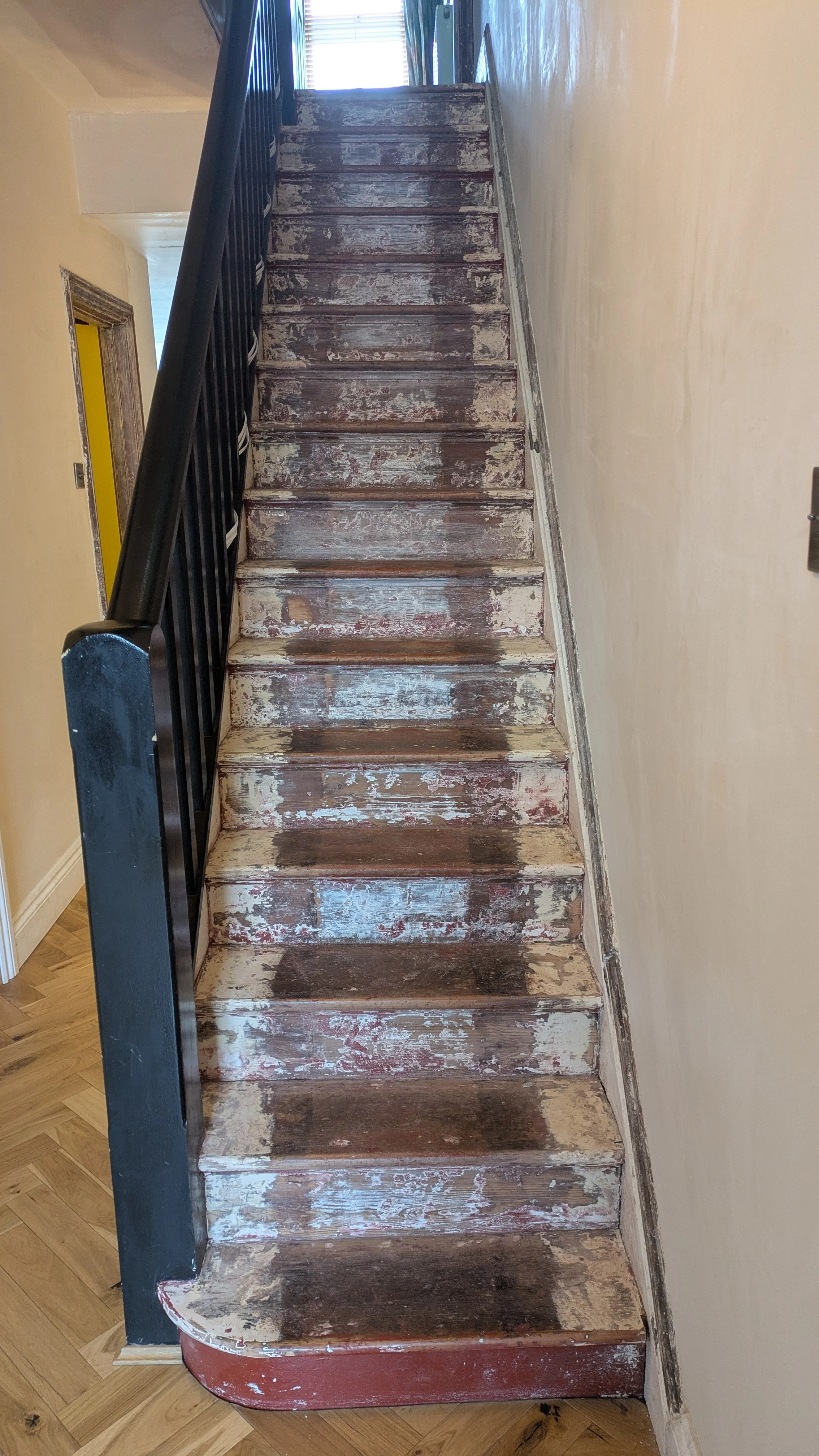 Old, worn wooden staircase with peeling paint, black metal railing on the left, leading up to a doorway with blinds on a window at the top.