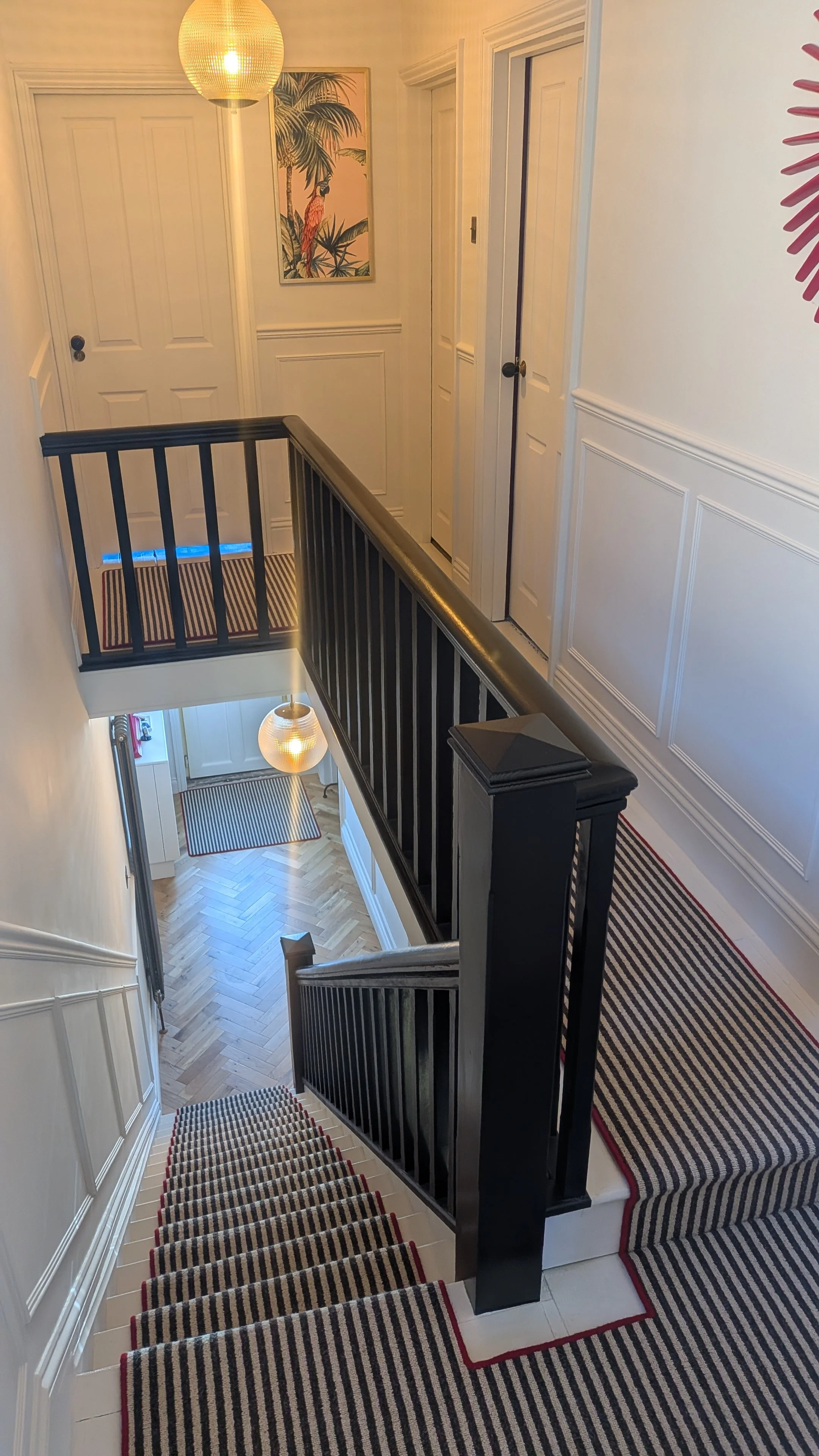 A staircase with black railings, striped carpet runners, and white walls, leading to a second floor with doors and a colorful tropical-themed artwork on the wall. A hanging light and a porch light are visible.