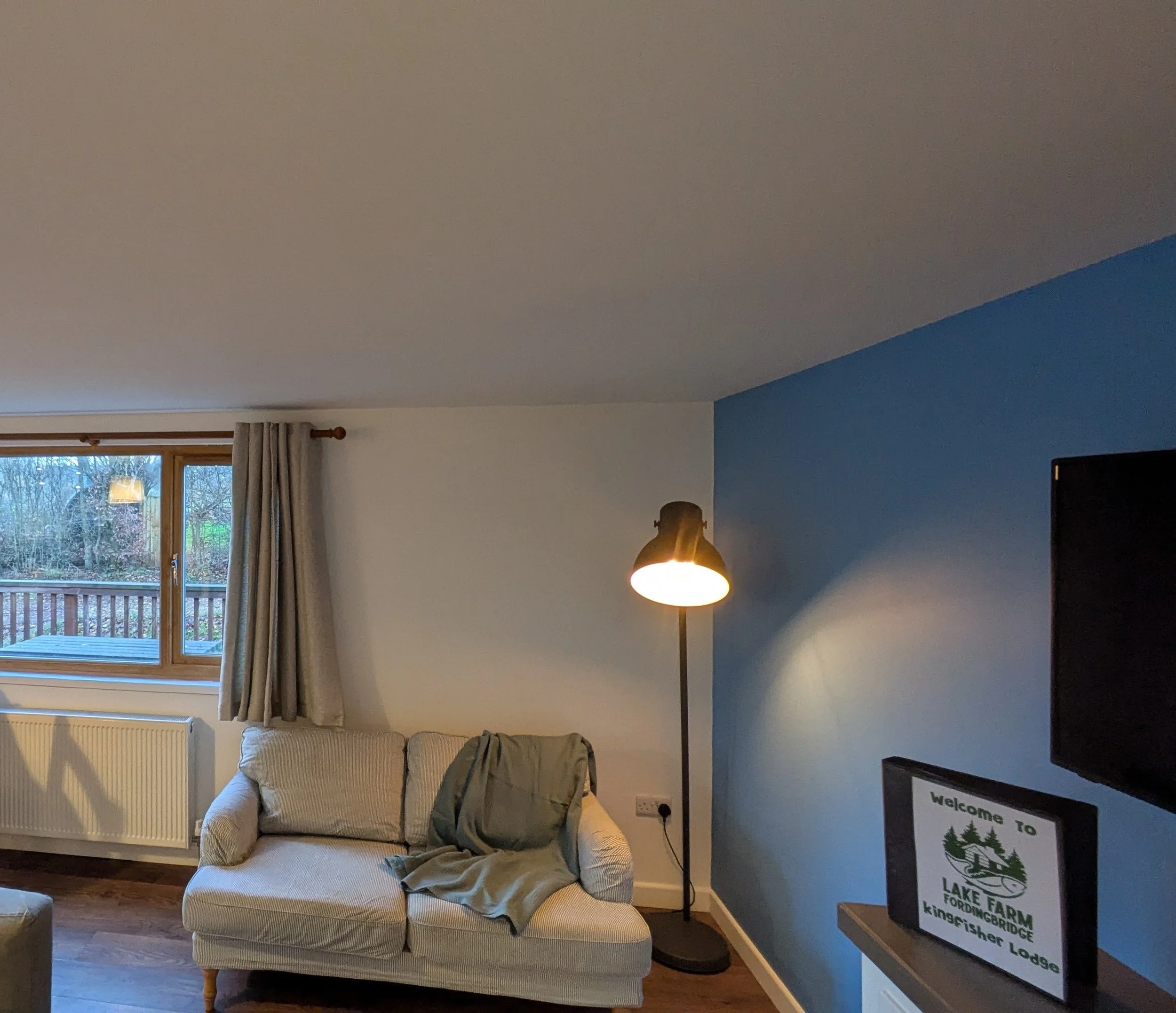 Living room with large window, beige sofa, green jacket on the sofa, floor lamp, and a framed sign that says "Welcome to Lake Farm, Kingfisher Lodge".