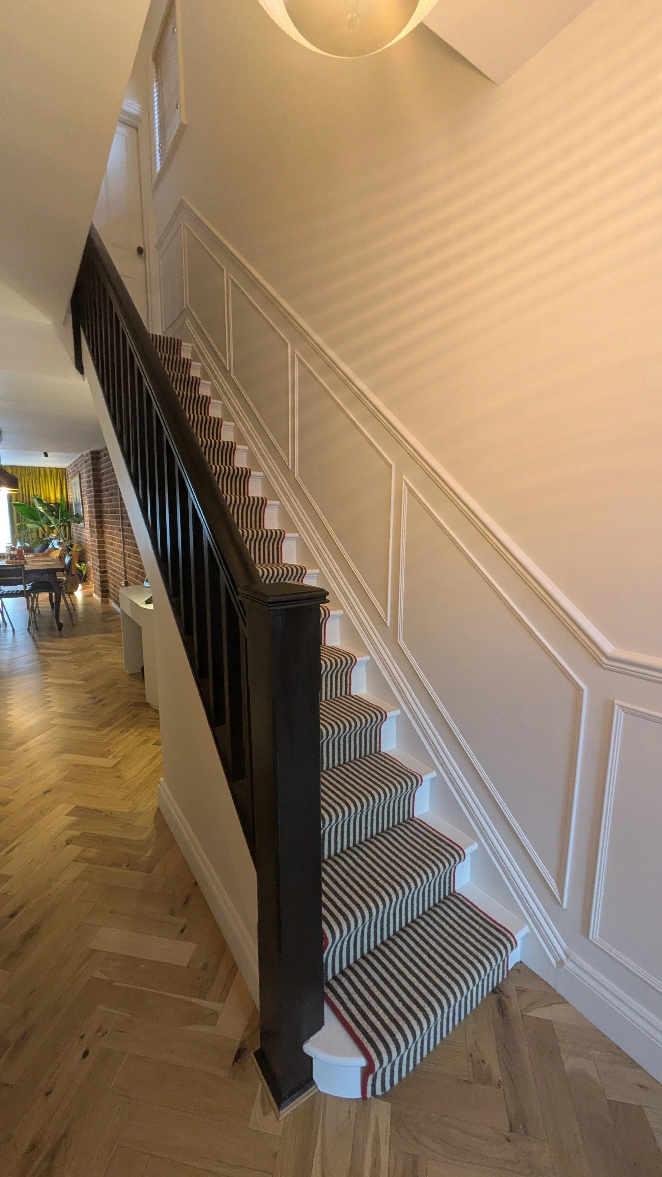 Interior view of a staircase with black railing, striped carpet runner, white wainscoting on the wall, and hardwood flooring, leading upstairs in a home.