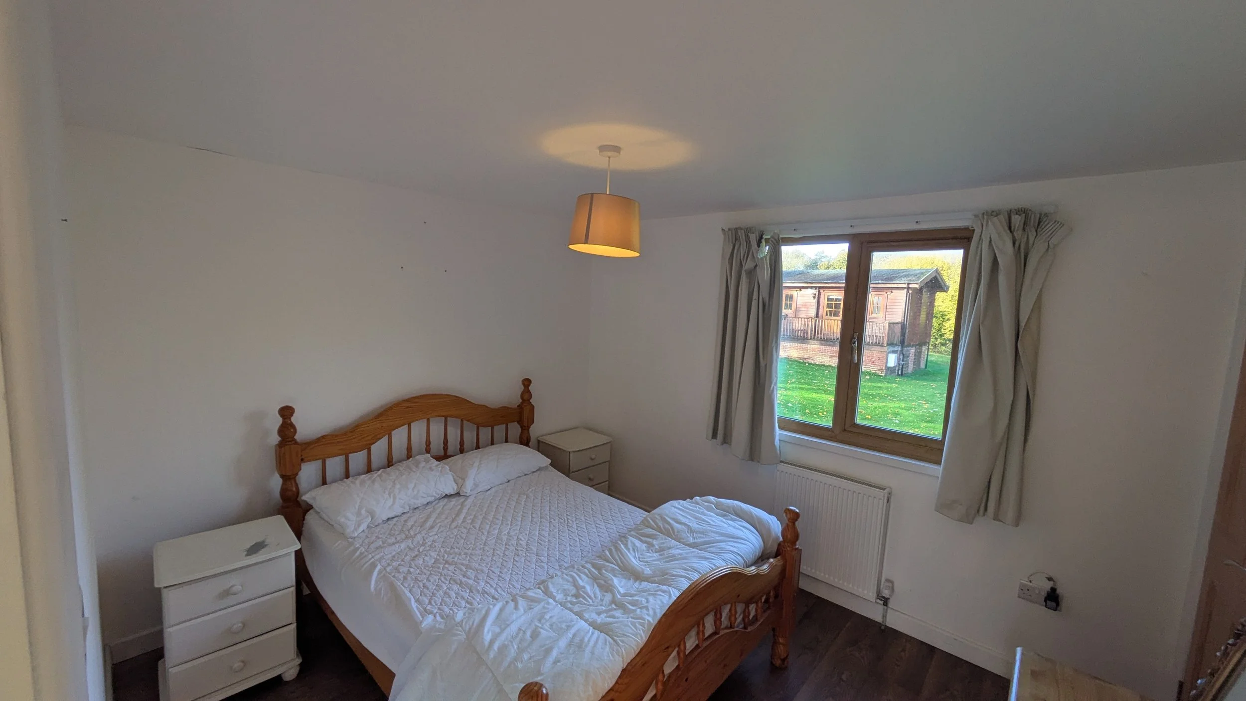 A bedroom with white walls, a wooden bed with white bedding, a white nightstand, a window with beige curtains, and a view of a wooden building outside.