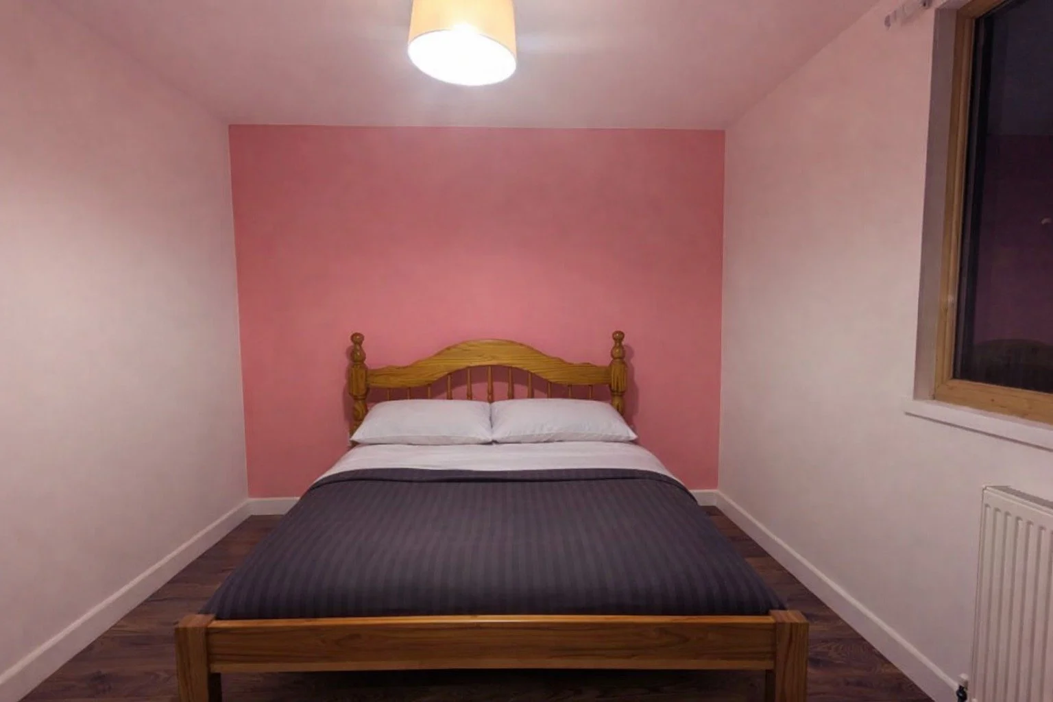 A bedroom with a bed, white pillows, a pink accent wall, and a window with a wooden frame.