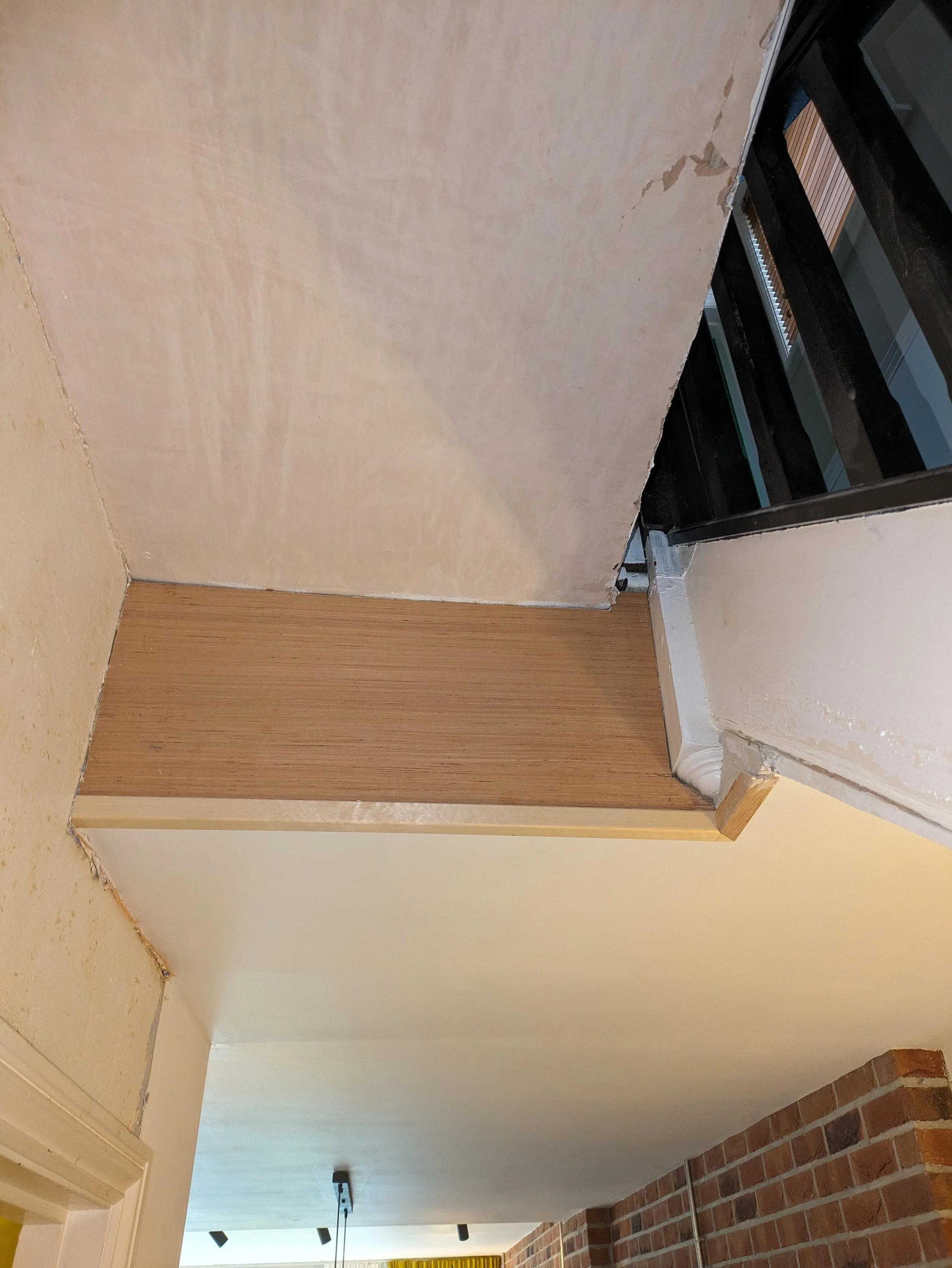 Interior view of a room under renovation, showing a partially finished parede with unpainted drywall, a corner with wood trim, and a brick floor. There are various construction elements, including a pipe and ceiling fixtures.