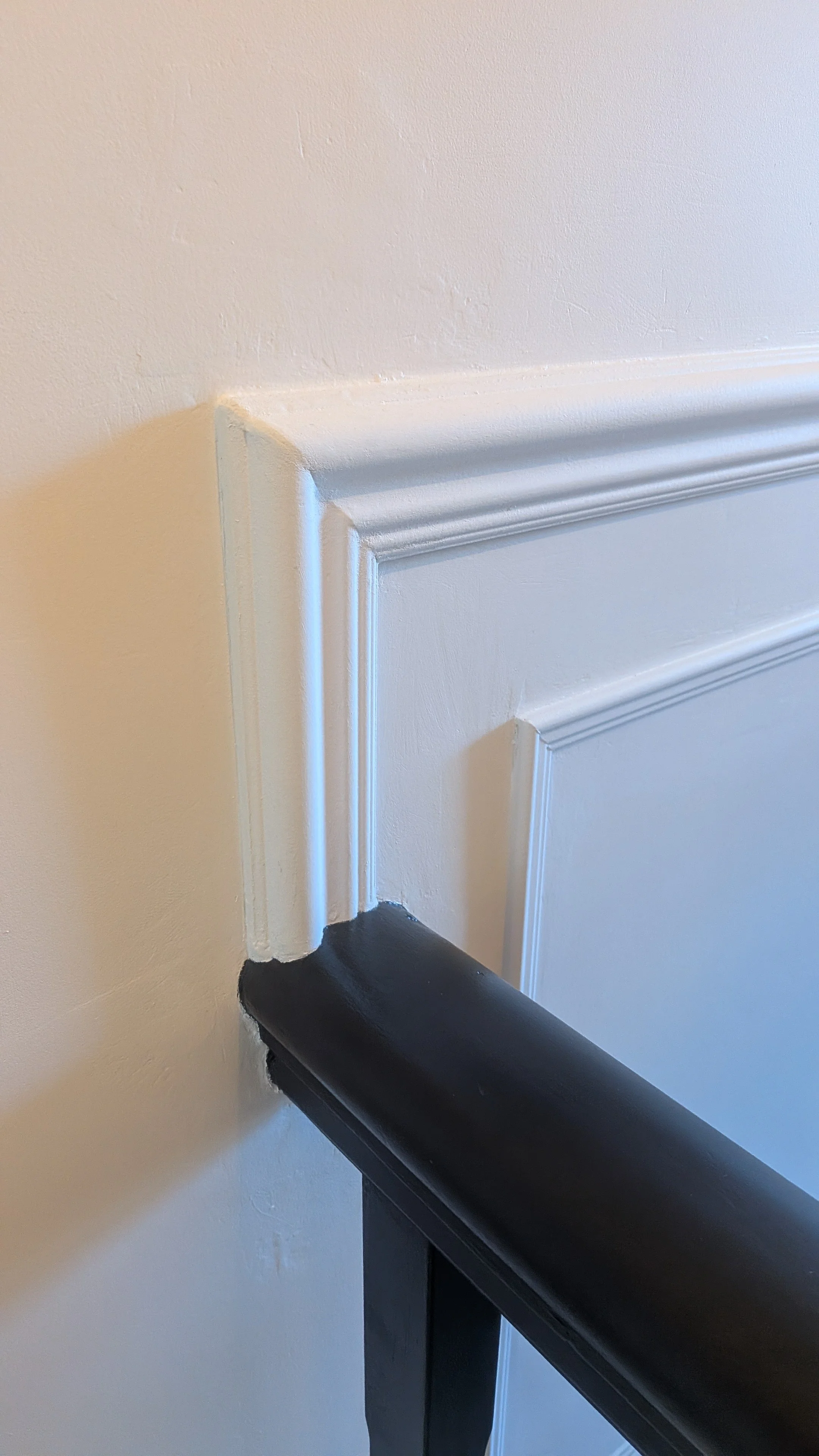 Close-up of a corner of a wall with white decorative molding and a black handrail at the bottom.
