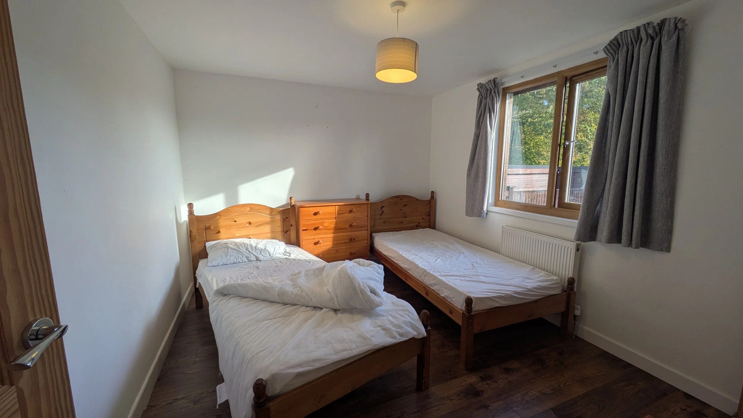 A bedroom with two single beds made of wood, one with white bedding and the other with a white sheet, facing a large window with gray curtains. There is a wooden dresser between the beds and a radiator beneath the window. The room has white walls and a ceiling light fixture.